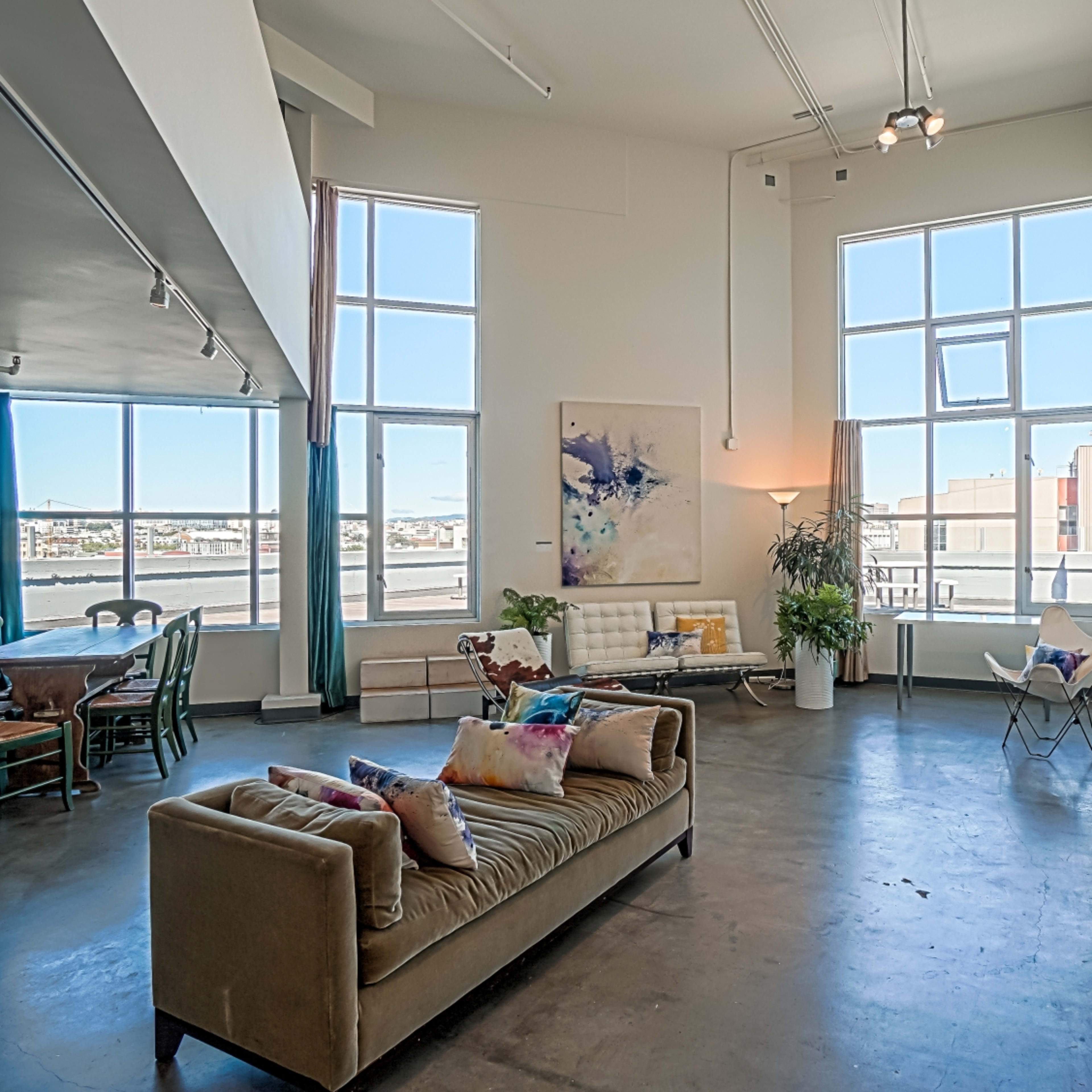 A spacious living area features large windows, a sofa with colorful pillows, a dining table, and abstract artwork on the walls.