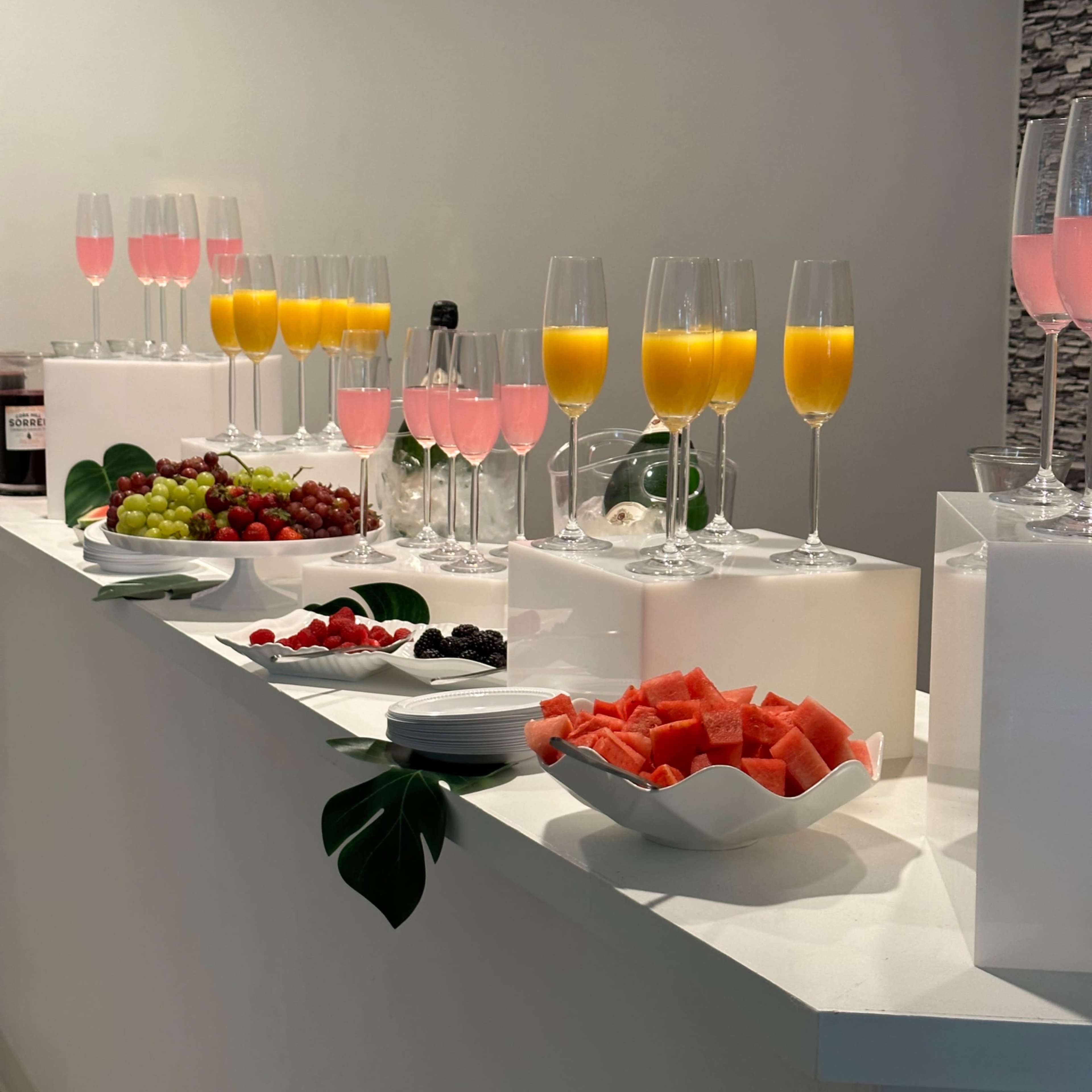 A long white countertop is adorned with glasses of pink and orange beverages, along with bowls of fresh fruit including grapes, strawberries, and watermelon.