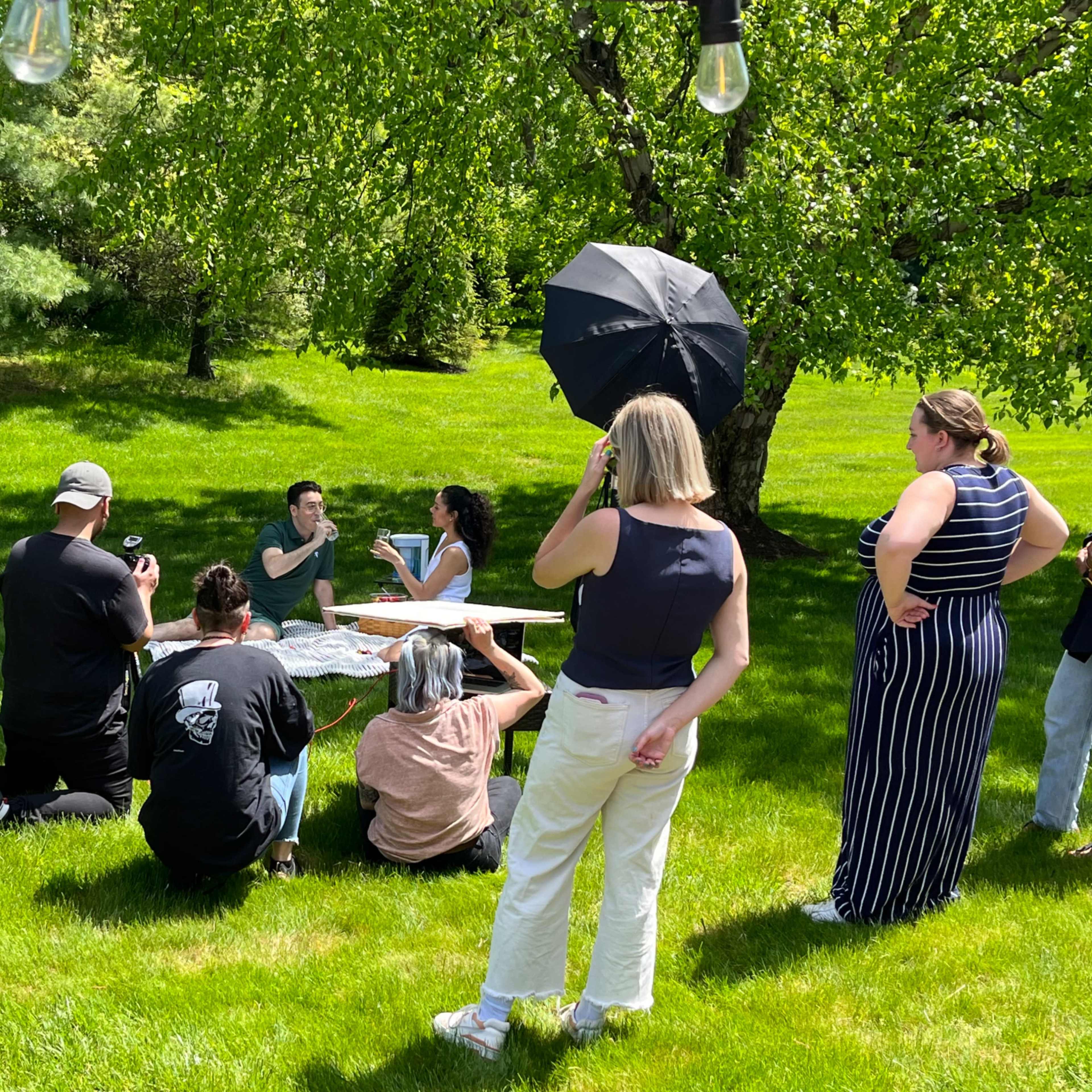 A group of people is gathered around a picnic table outdoors, with some taking photos while others are engaged in conversation under a sunny sky.