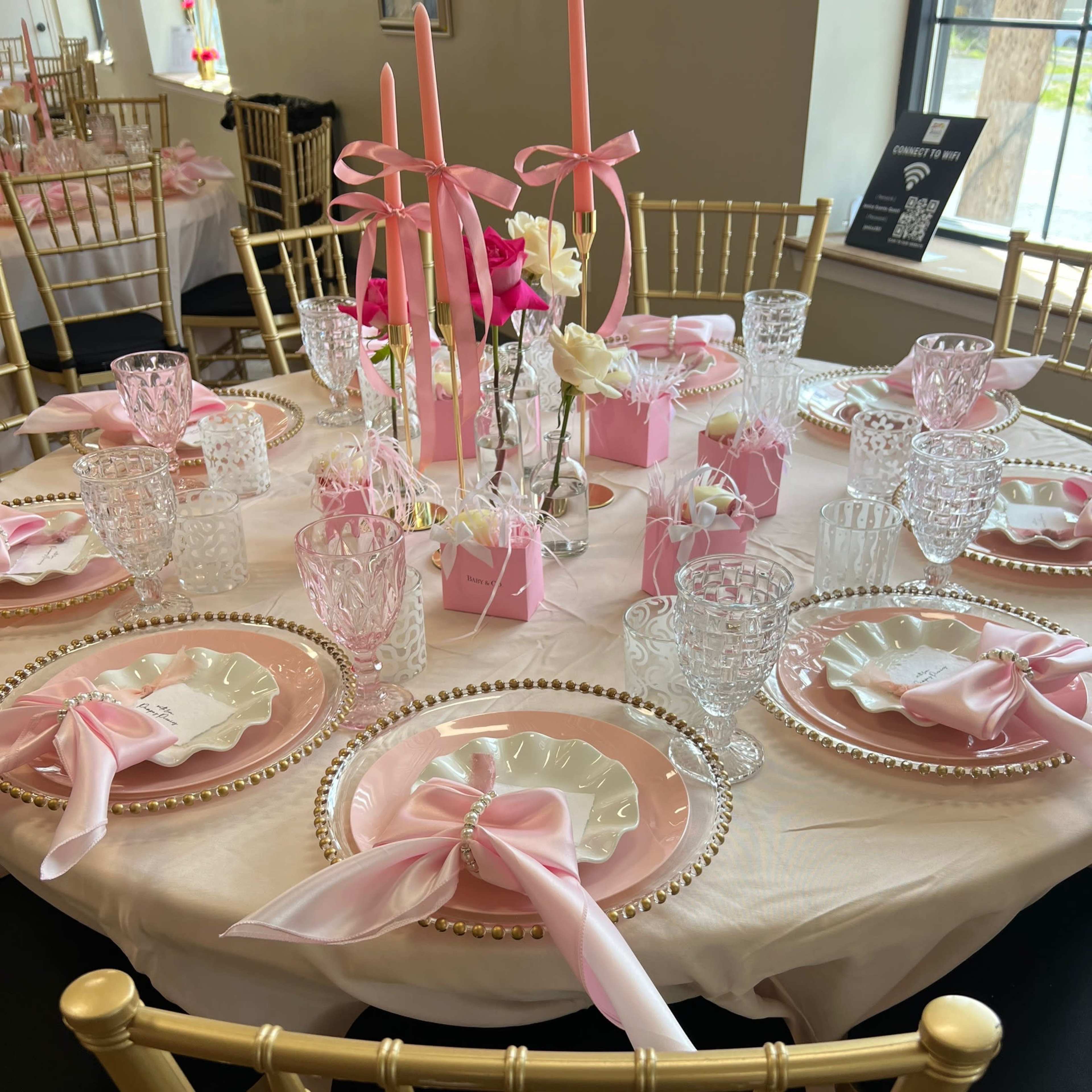 A beautifully set dining table features pink and white tableware, elegant glassware, and decorative floral arrangements.