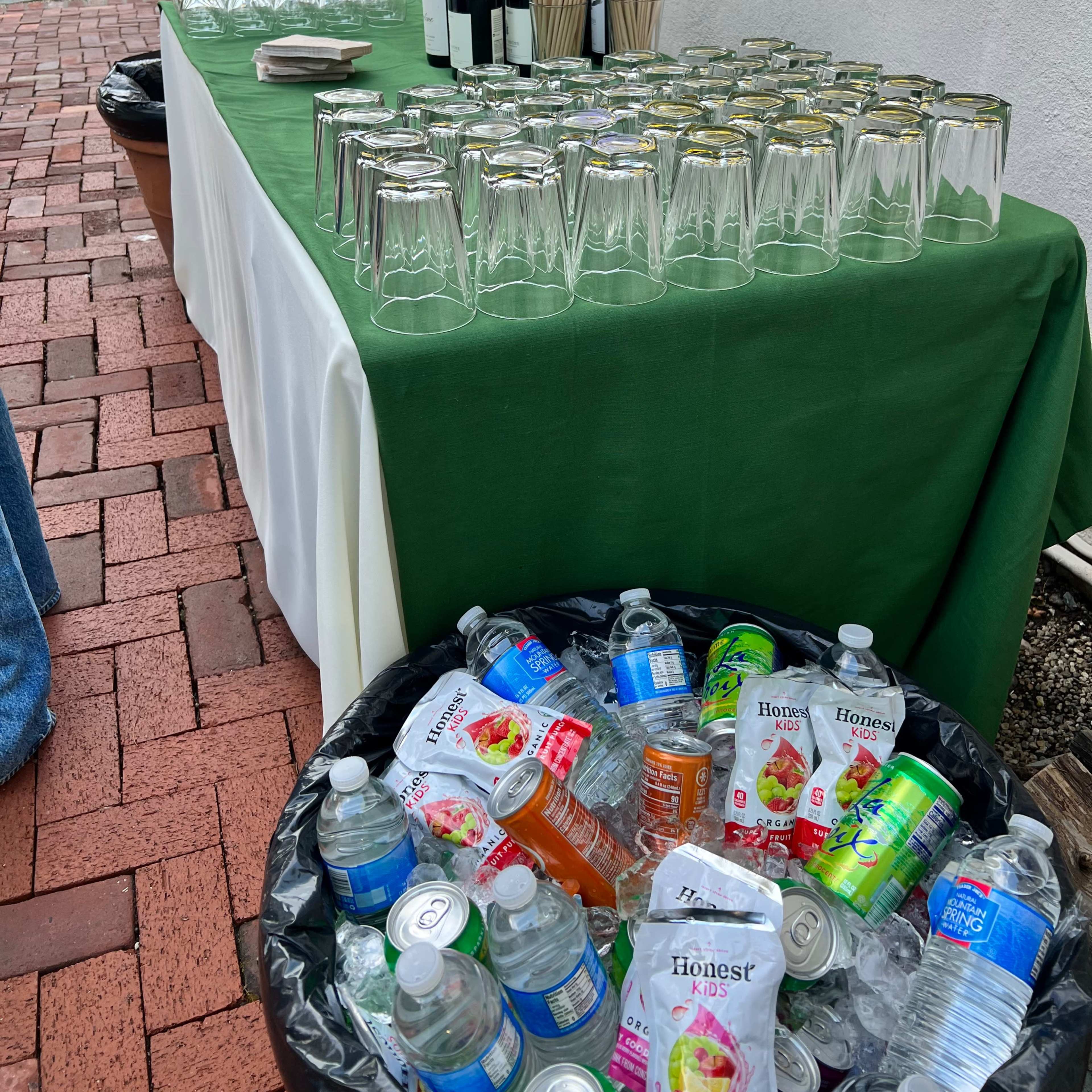 A table set with glasses, wine bottles, and drink straws is positioned beside a bin overflowing with empty beverage containers.