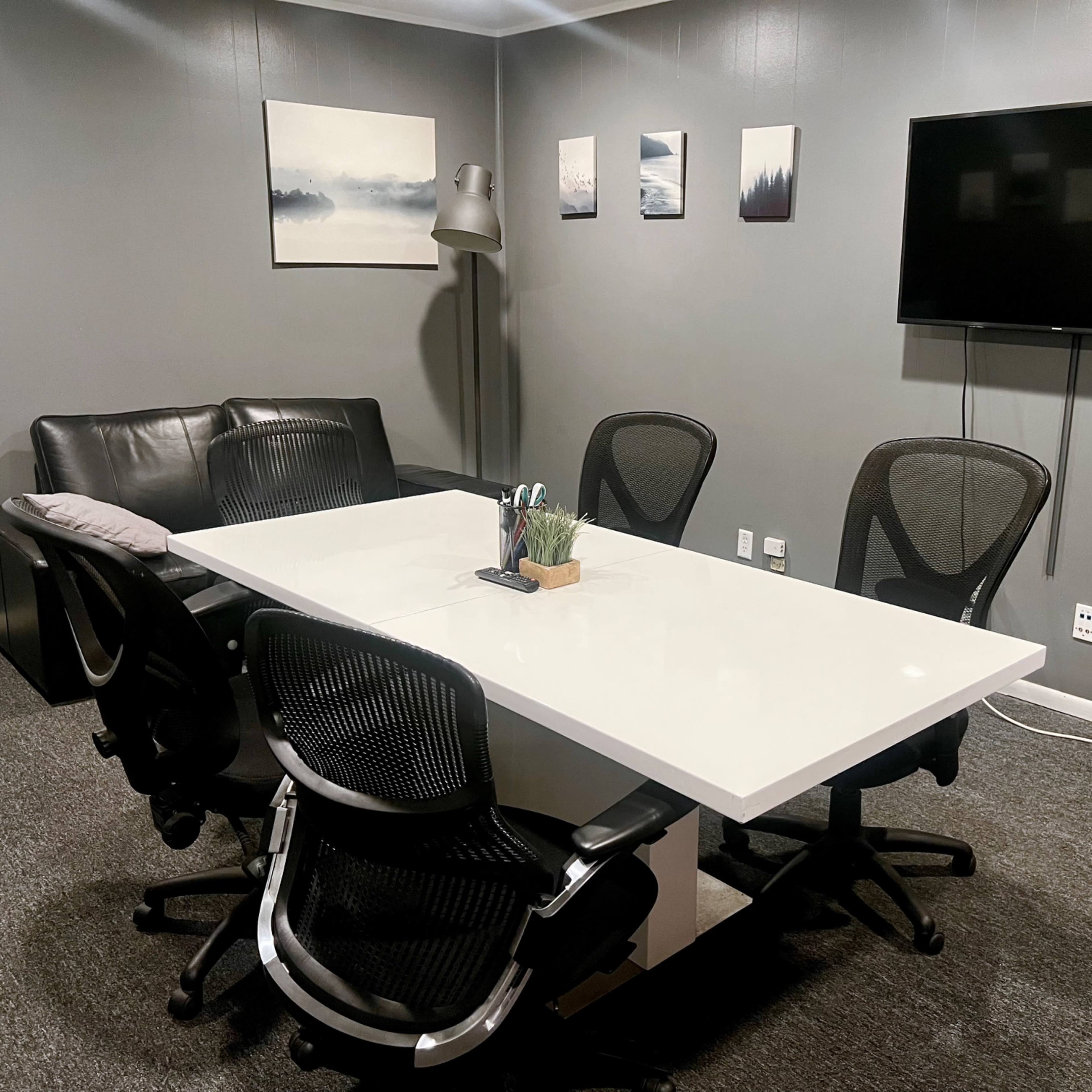 A modern conference room with a rectangular white table surrounded by four black office chairs, a sofa, a lamp, and a wall-mounted television.