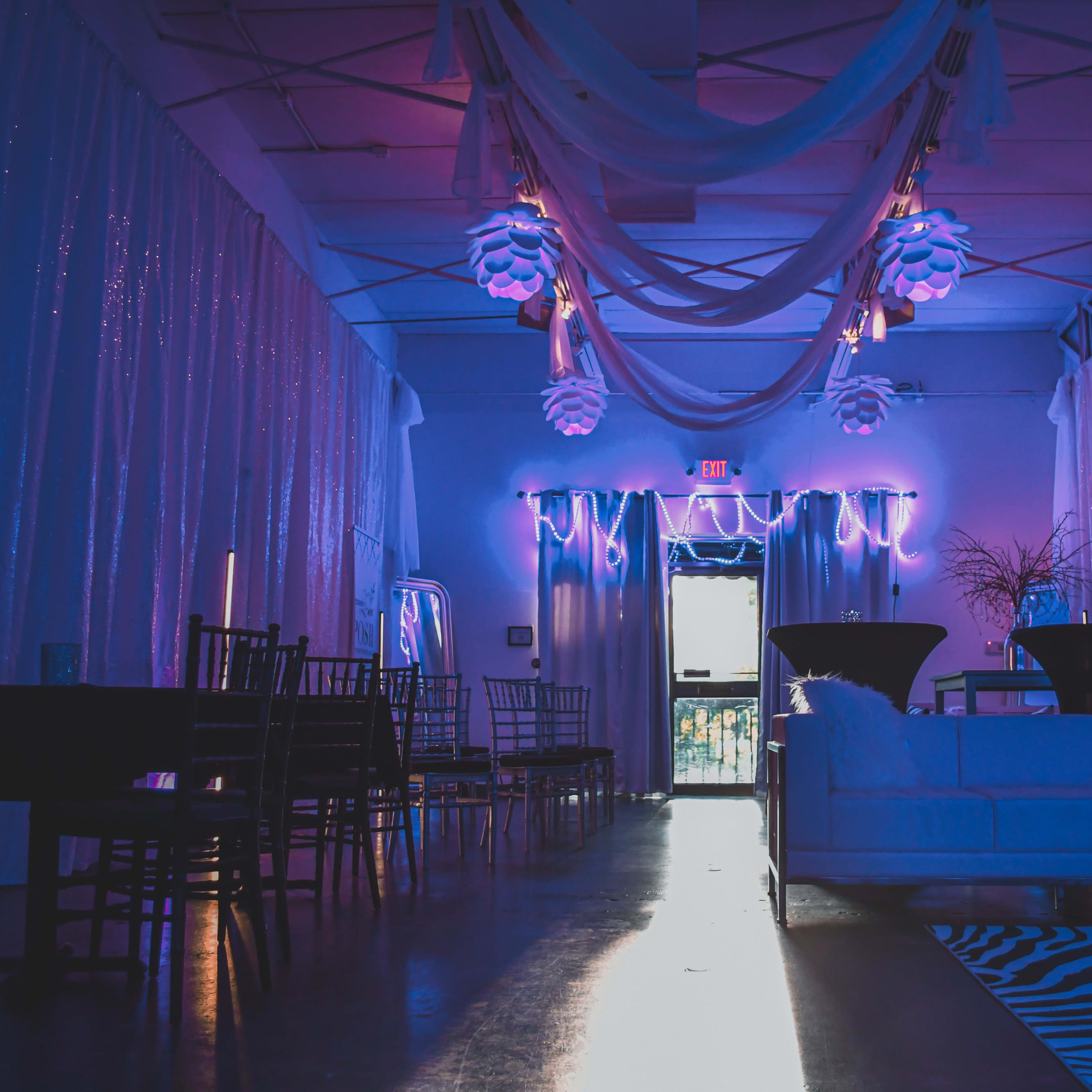 A decorated indoor event space with draped fabric, purple lighting, and a seating area featuring a white couch and black chairs.
