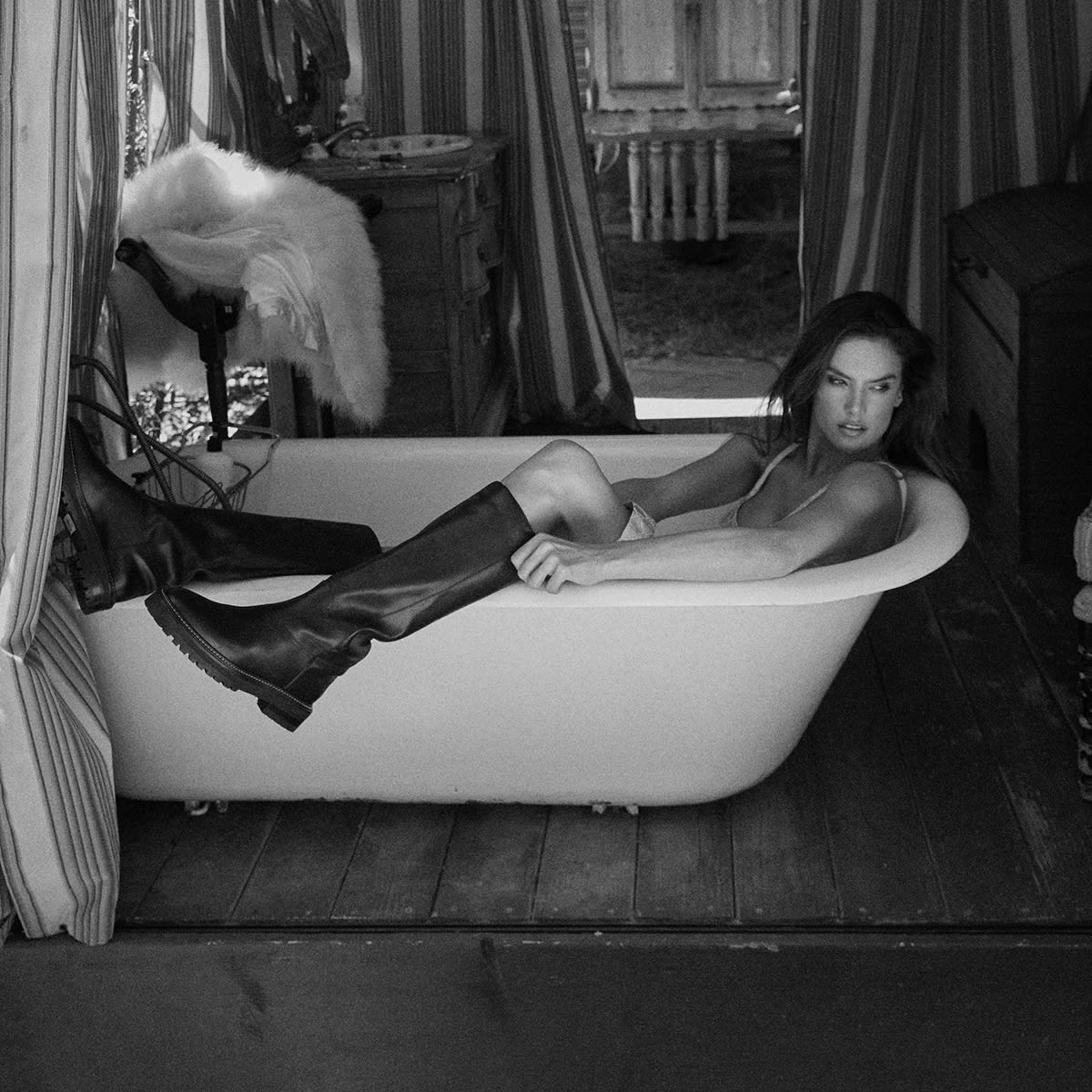 A woman in knee-high boots reclines in a vintage bathtub inside a room with striped curtains and wooden furnishings.