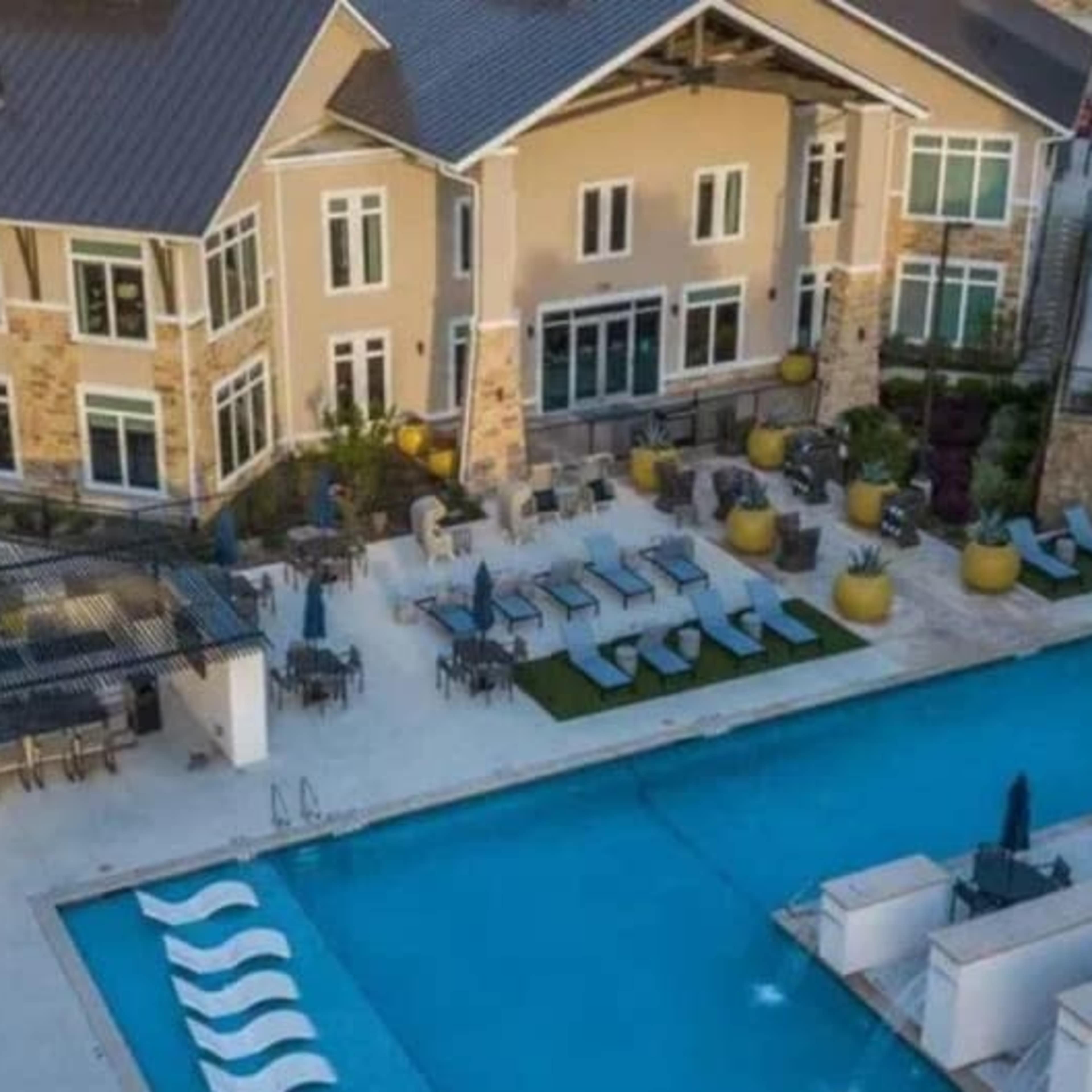 A modern residential building adjacent to a large outdoor pool surrounded by lounge chairs and landscaped areas.
