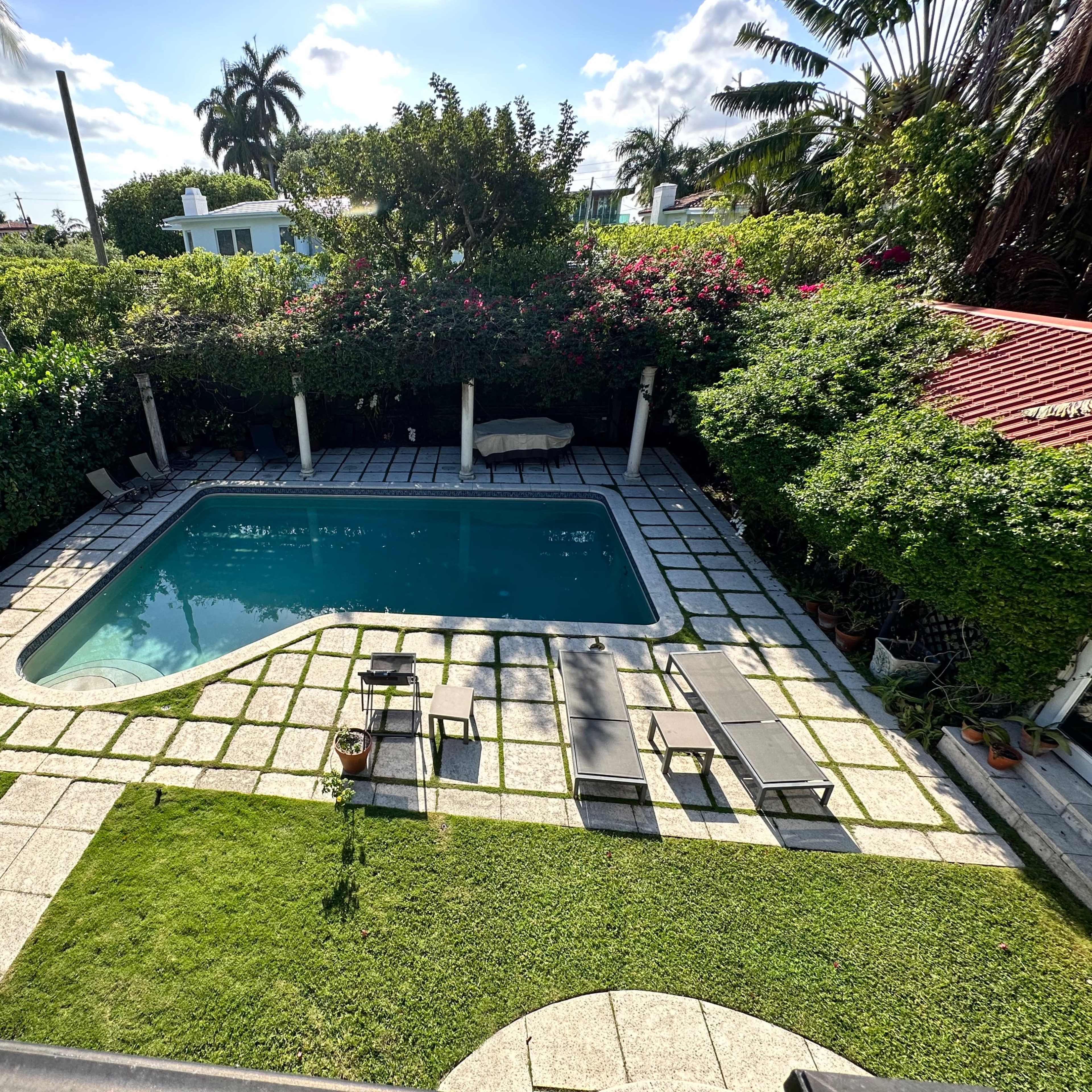A rectangular swimming pool surrounded by green foliage and patio furniture in a landscaped yard.