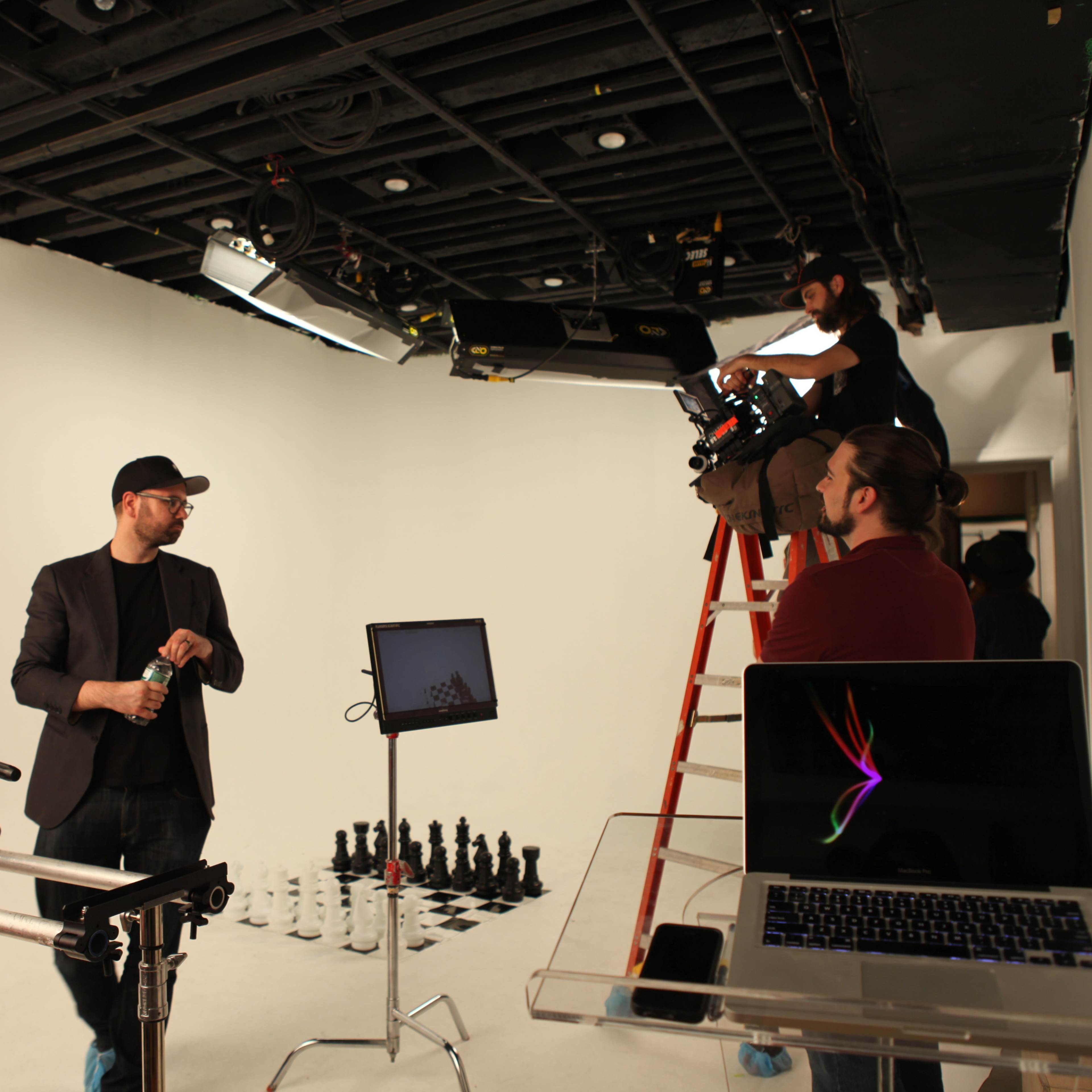 A film crew is preparing a set in a studio with a white backdrop, featuring a chessboard and equipment including cameras and a laptop.