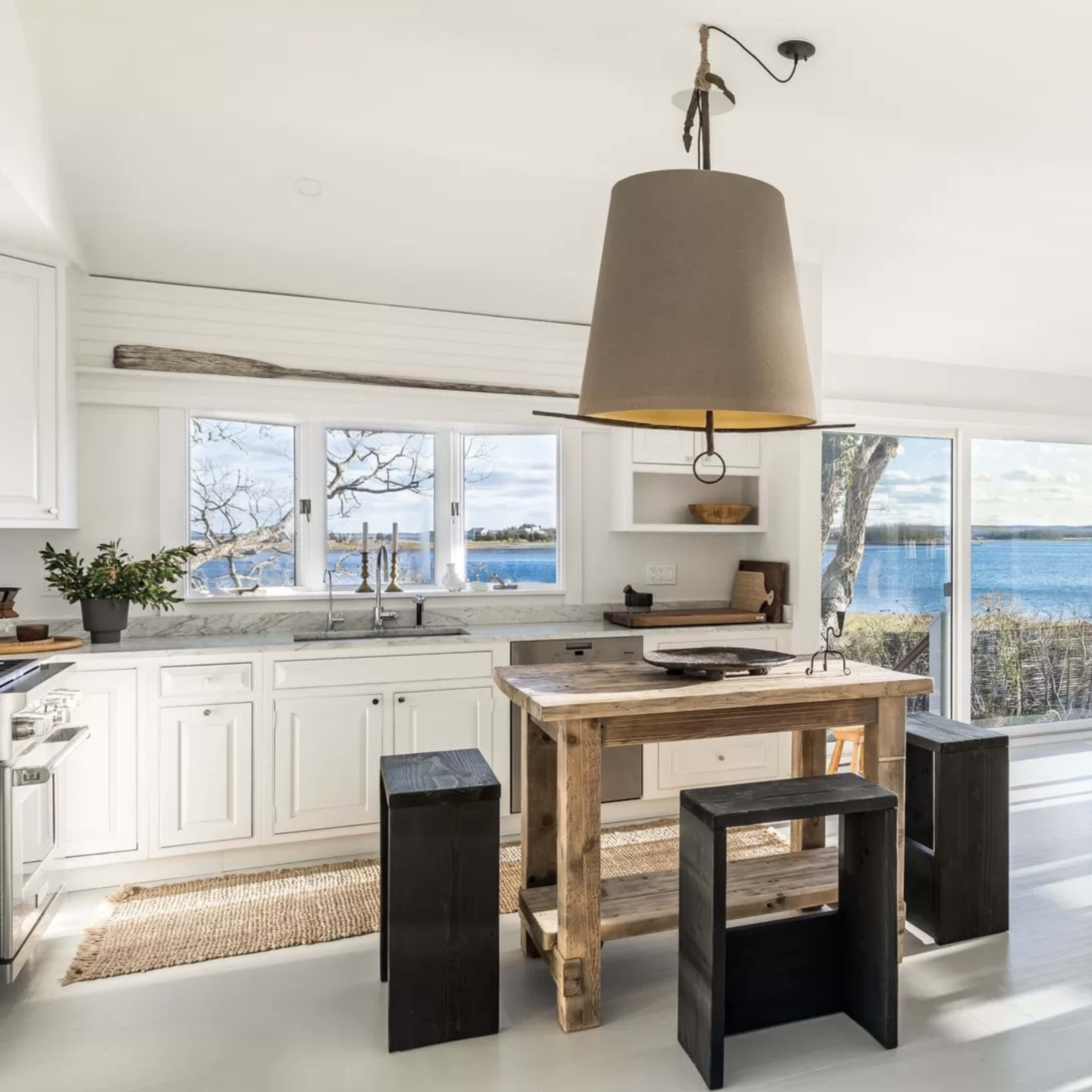 A bright kitchen features a wooden island with stools, large windows showcasing a view of the water, and a dining area with a table and chairs.