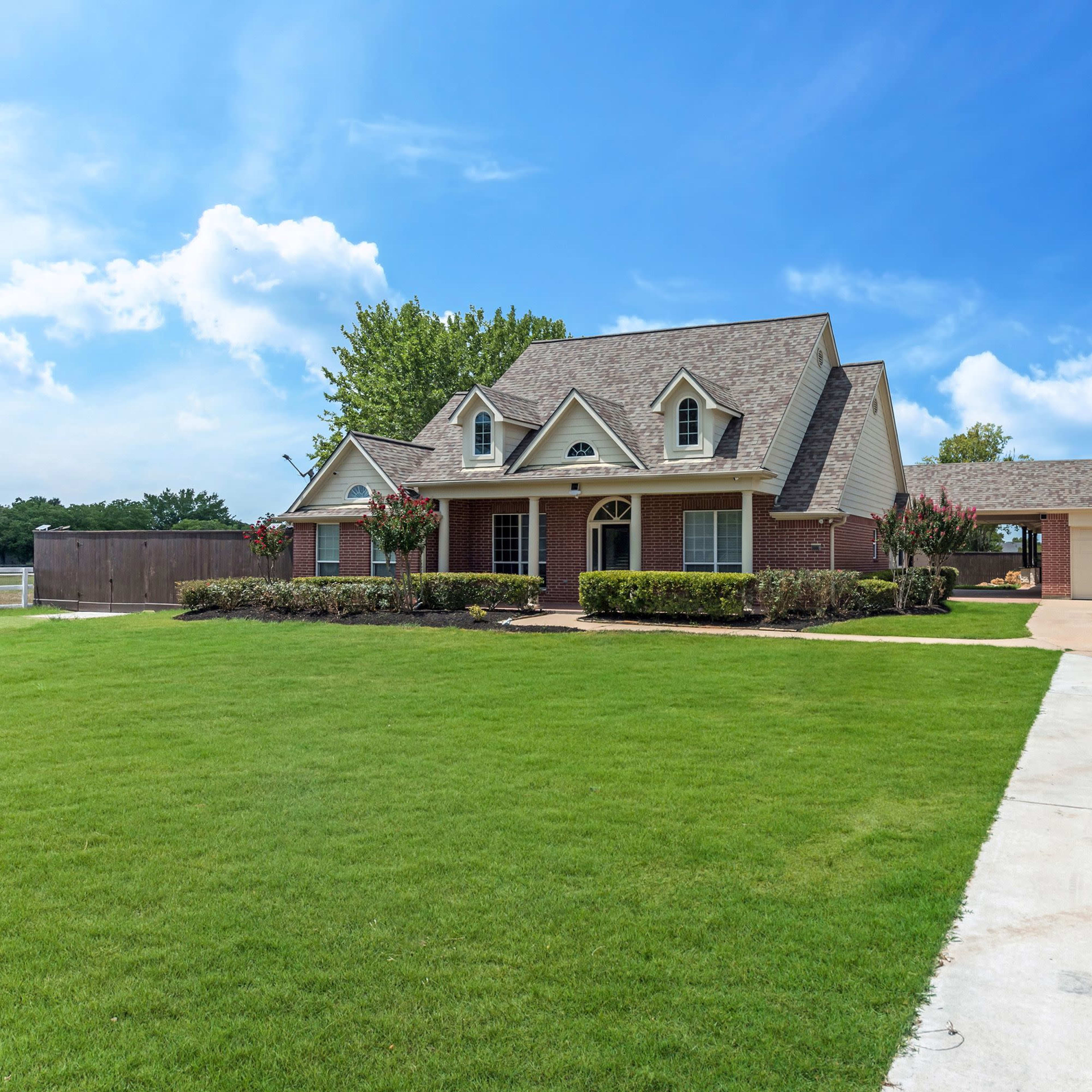 A brick house with a sloped roof and a garage sits on a spacious green lawn under a partly cloudy sky.