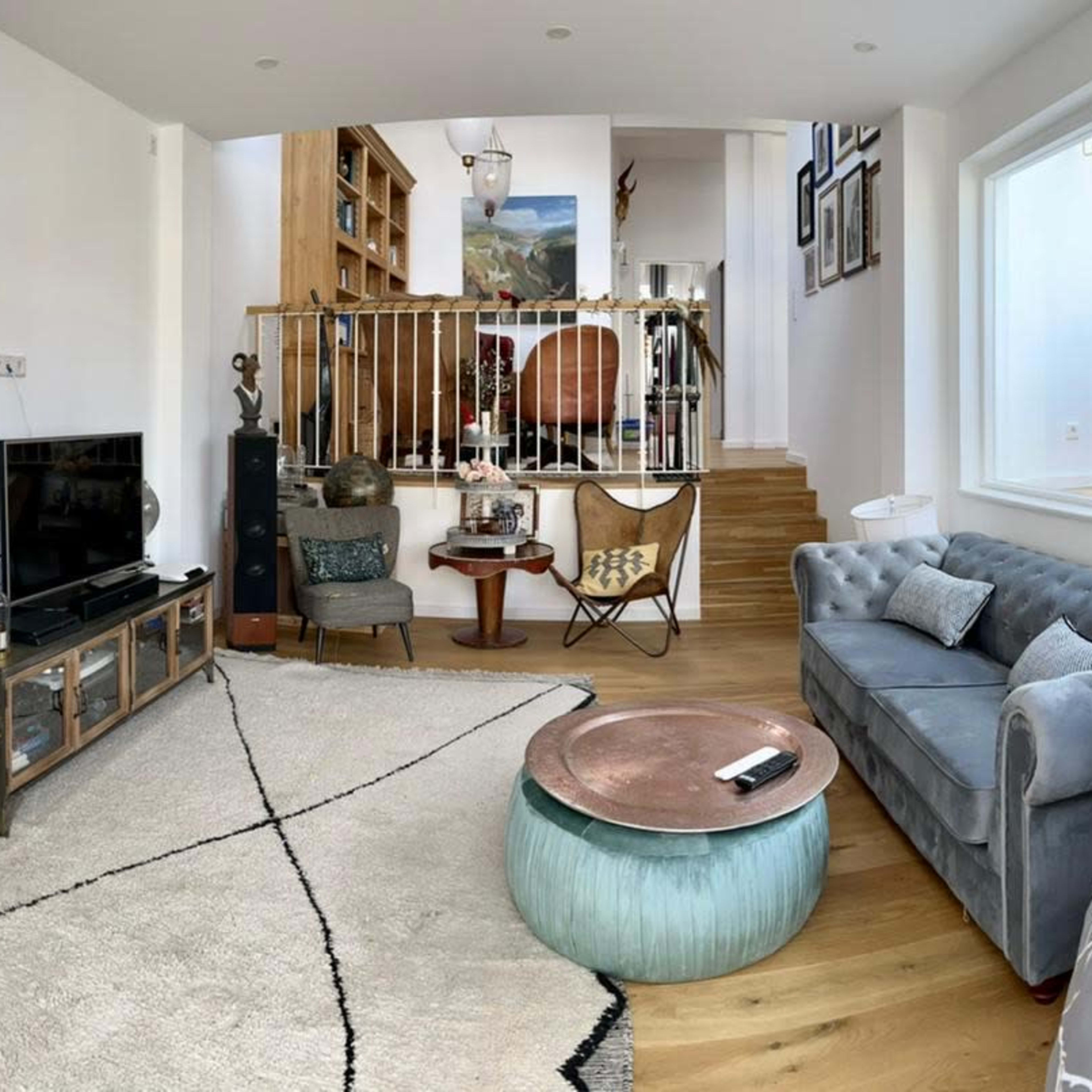 A stylish living room with a mix of furniture, including a blue sofa, a rattan chair, and a green coffee table, set within a bright, open space that features a staircase and wooden flooring.