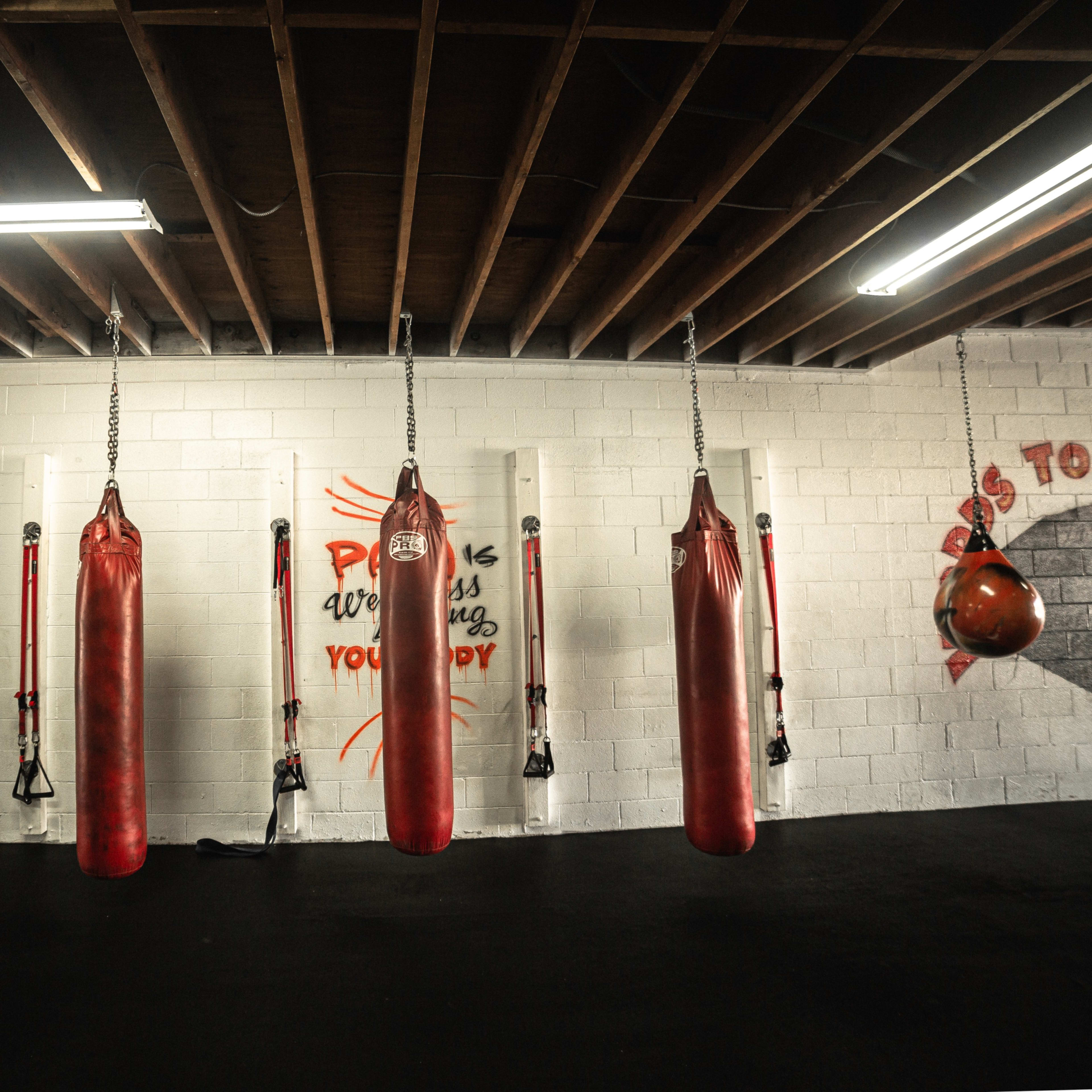 A boxing gym with several red heavy bags hanging from the ceiling, mounted on wall brackets, and motivational slogans painted on the wall.
