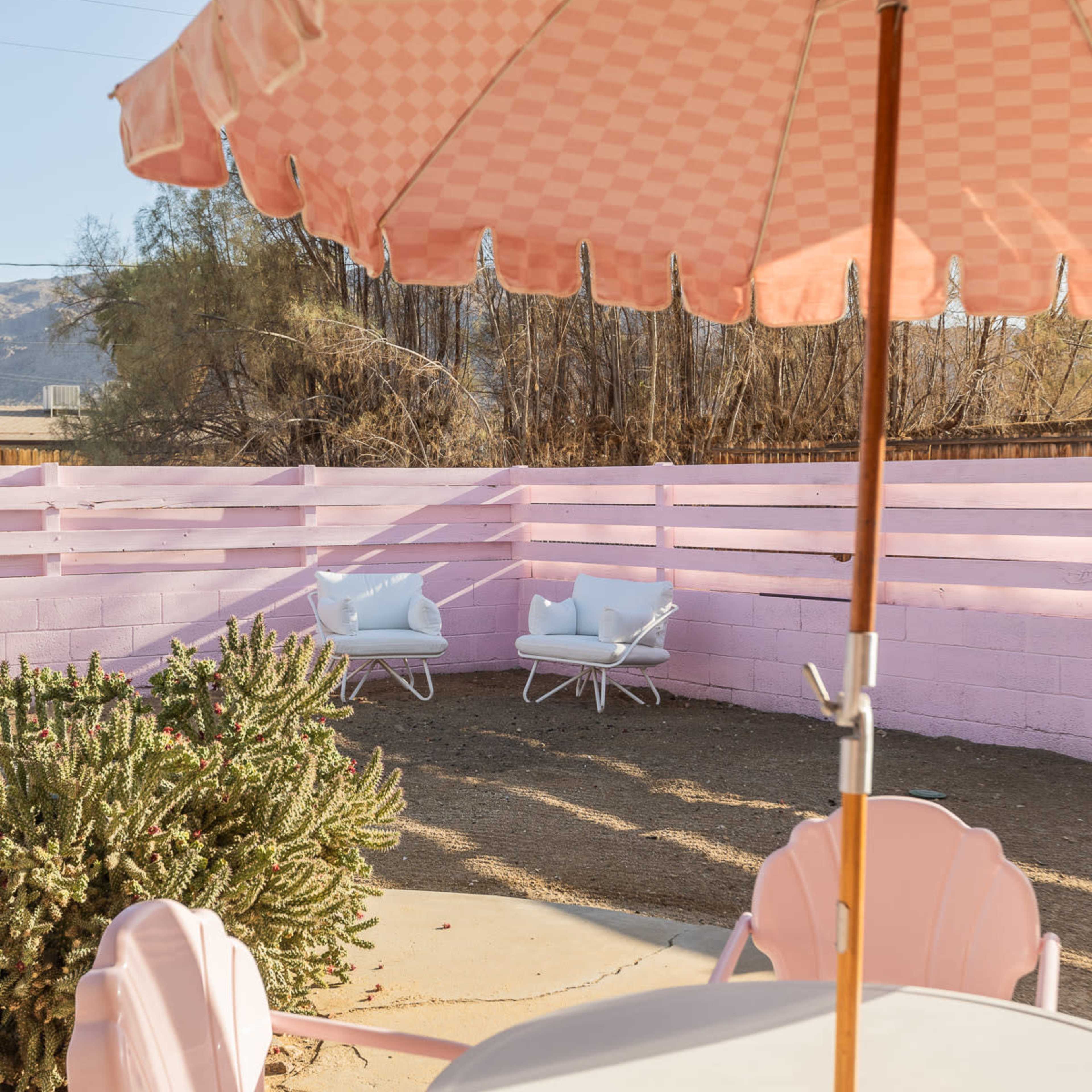 A pink umbrella shades a round table and chairs, overlooking a fenced area with two white armchairs and sparse vegetation.