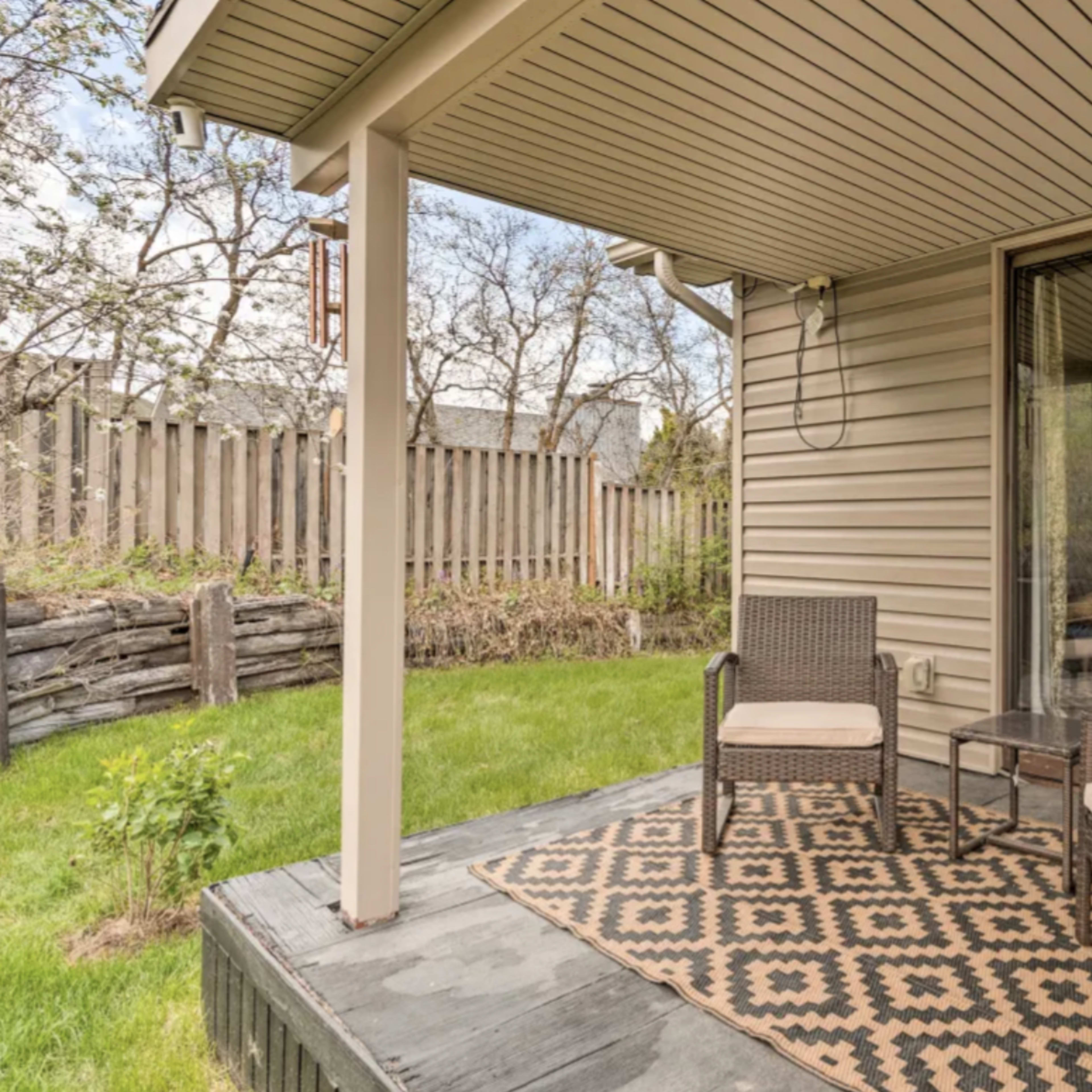 A covered patio features two chairs and a glass table on a patterned rug, with a grassy area and trees in the background.