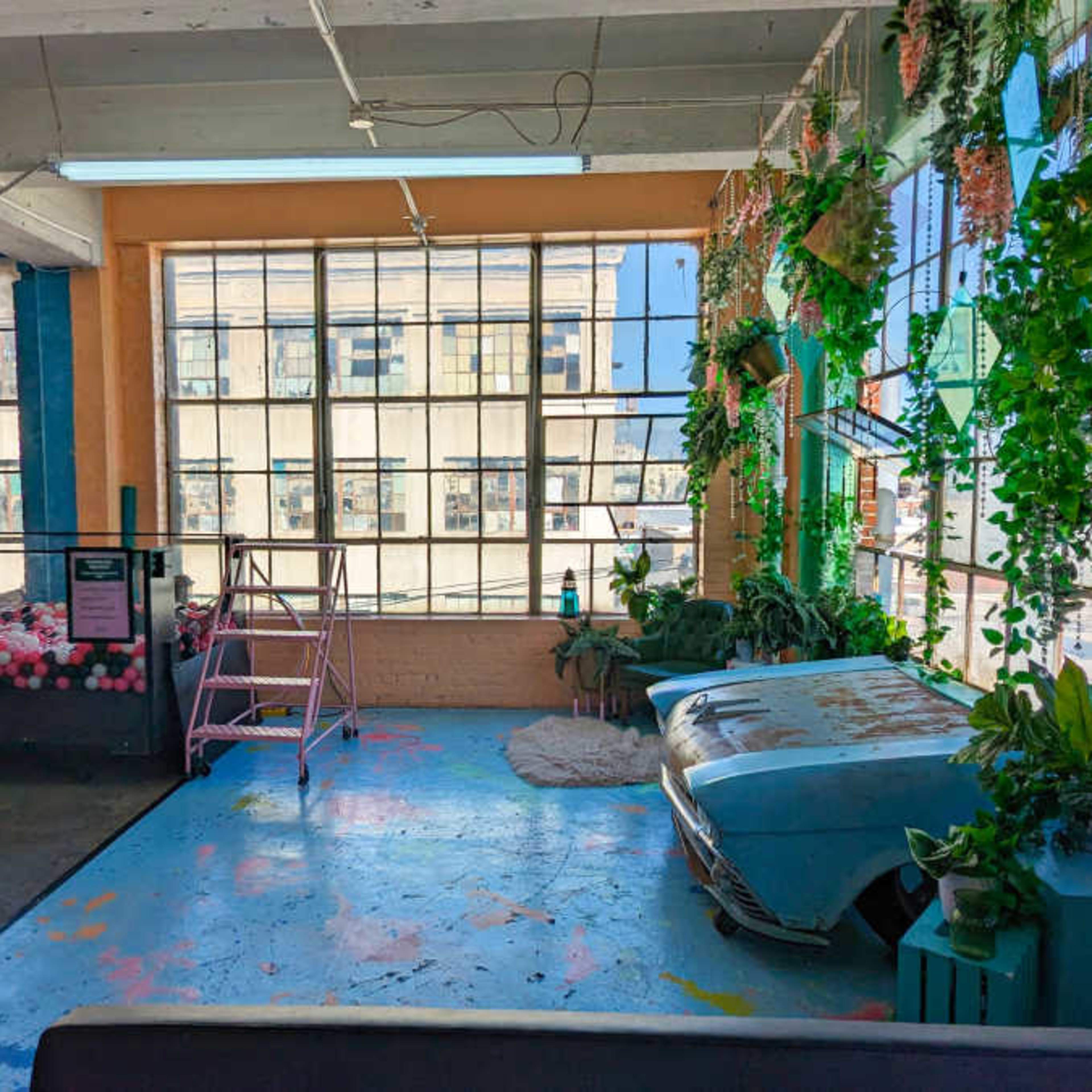 An industrial-style studio with large windows, colorful walls, and various plants arranged throughout the space.