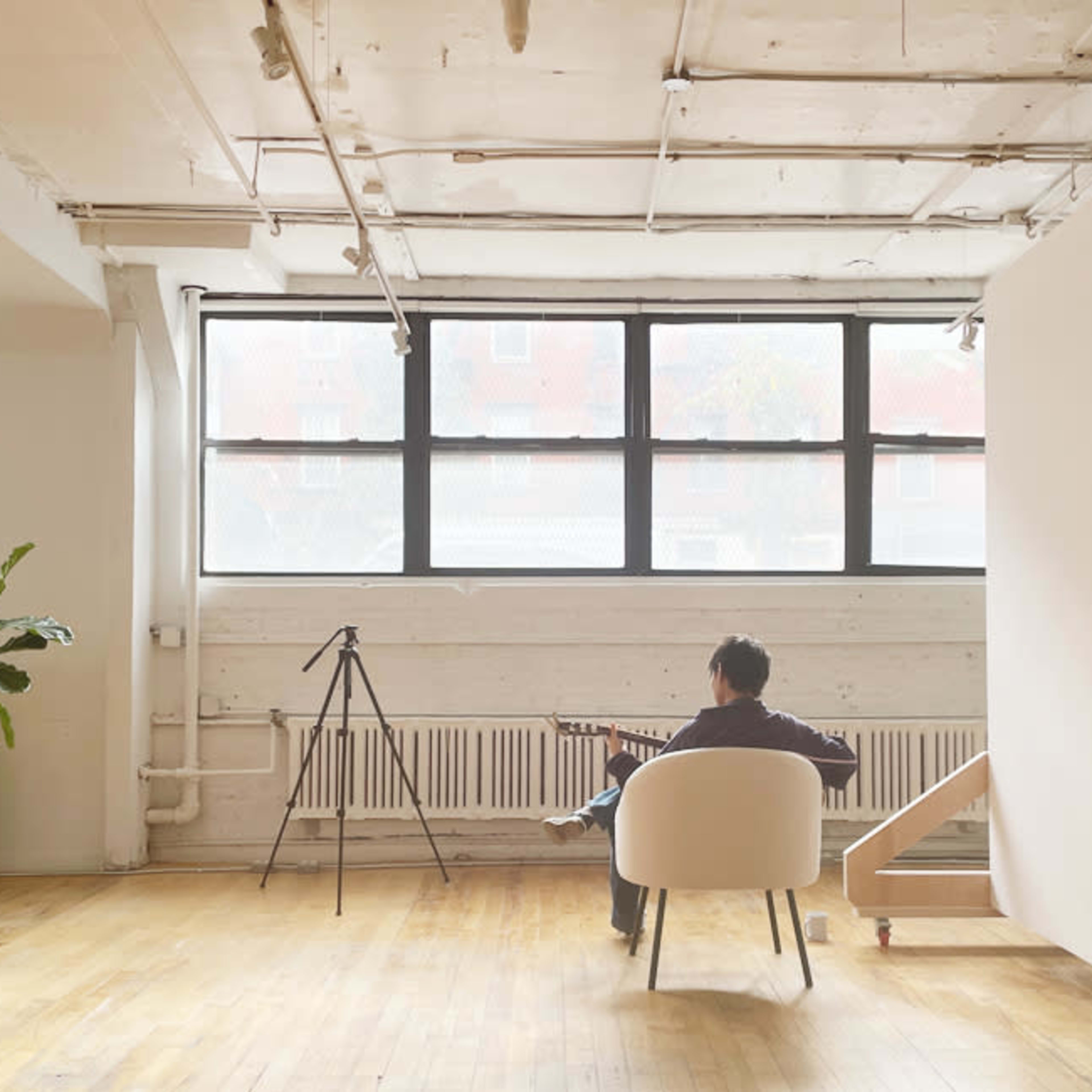 A person sits in a white chair in a spacious, minimalist room with large windows, a plant, and a tripod.