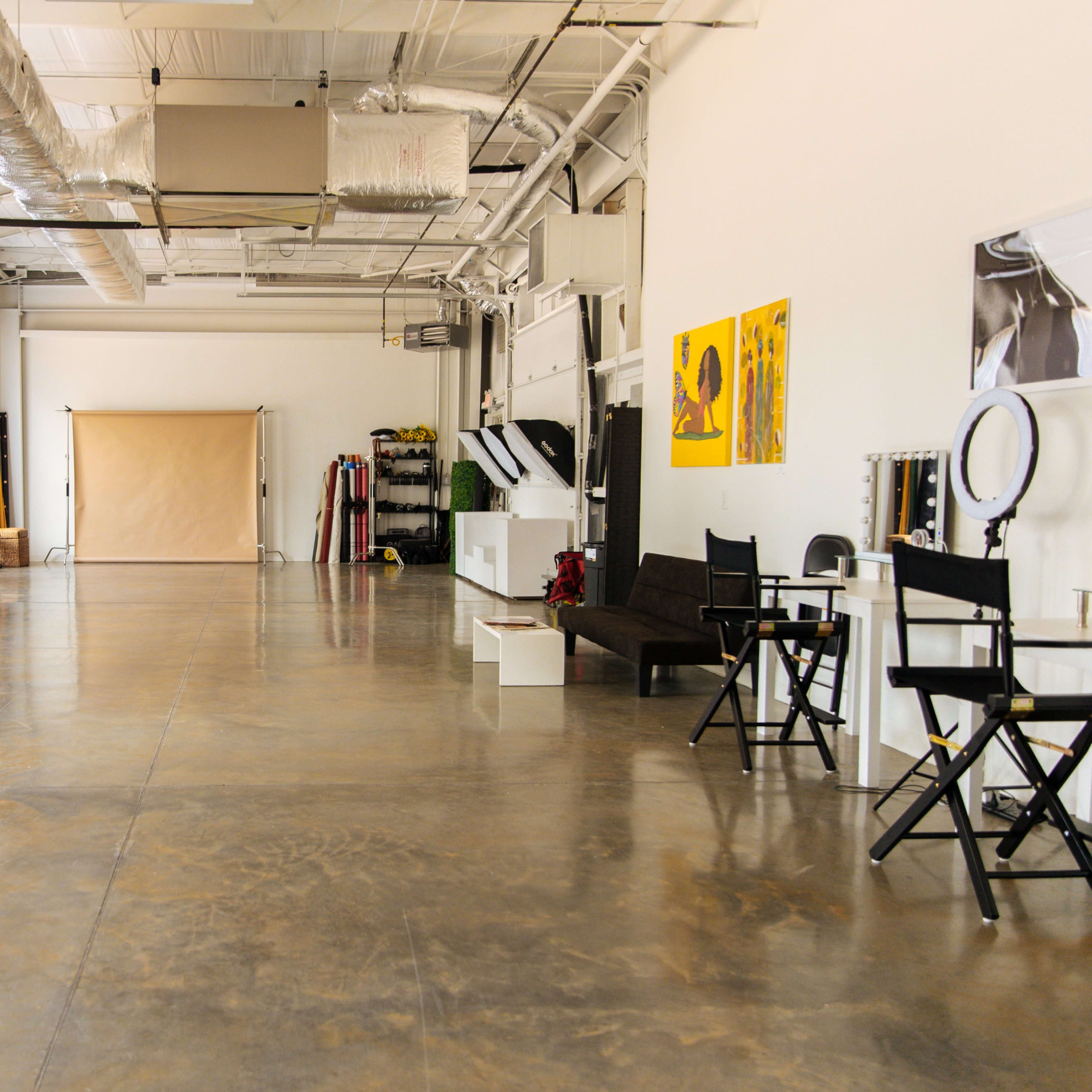 The interior of a spacious, well-lit studio features a polished concrete floor, large windows with sheer curtains, and various furniture pieces including a couch and director's chairs.