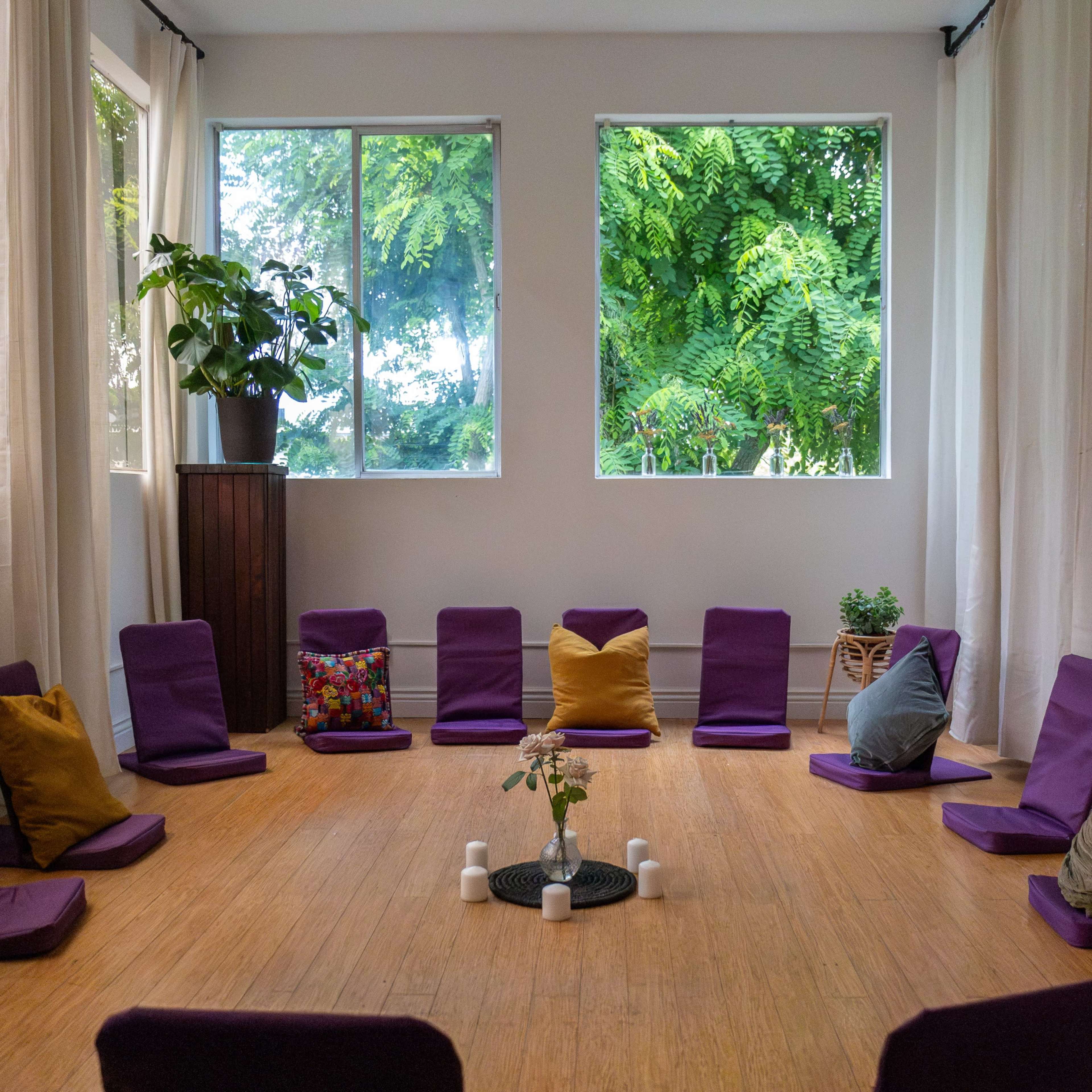 A circular arrangement of purple cushions is set up in a bright room with large windows overlooking greenery.