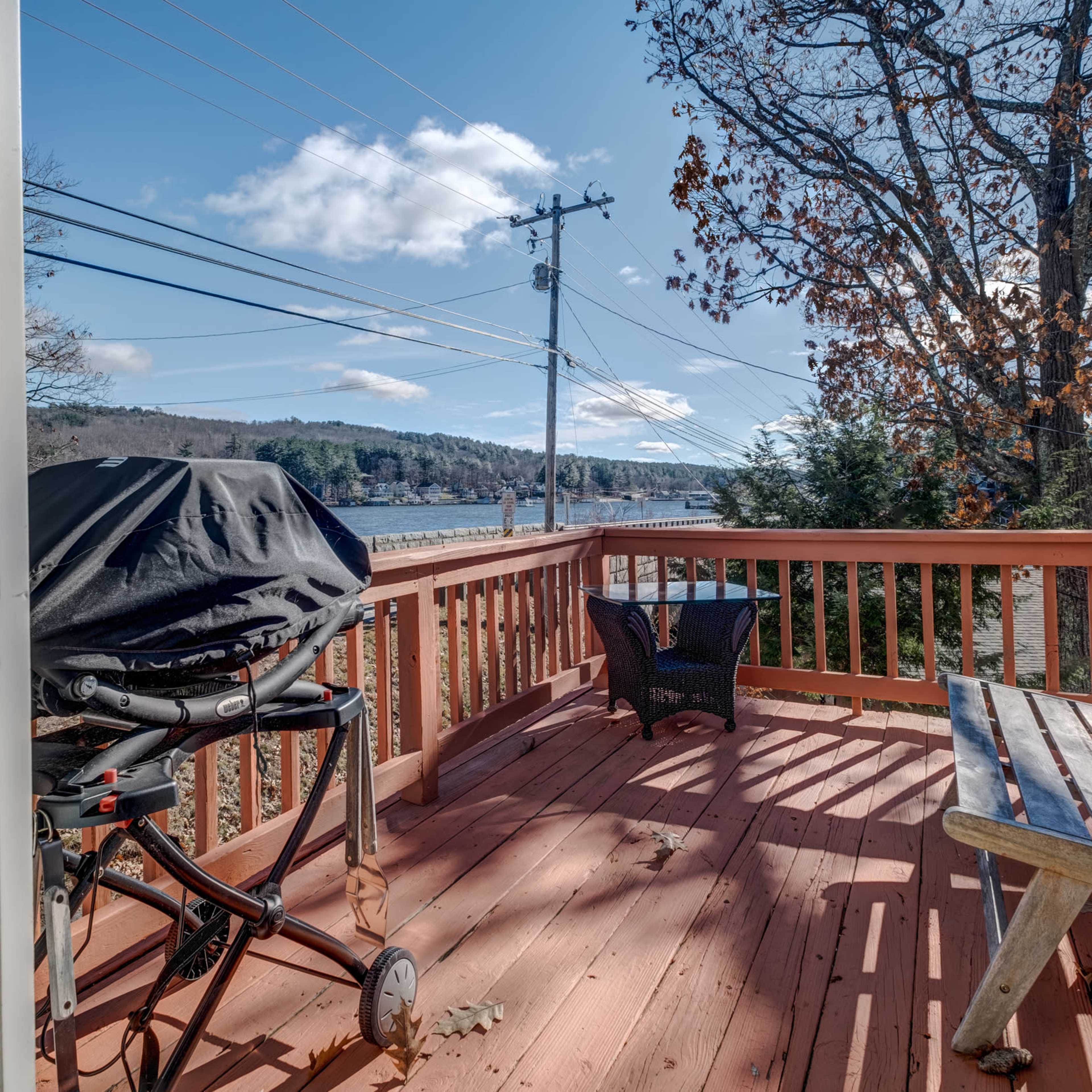 A wooden deck with a grill and a bench overlooks a lake and trees under a clear blue sky.