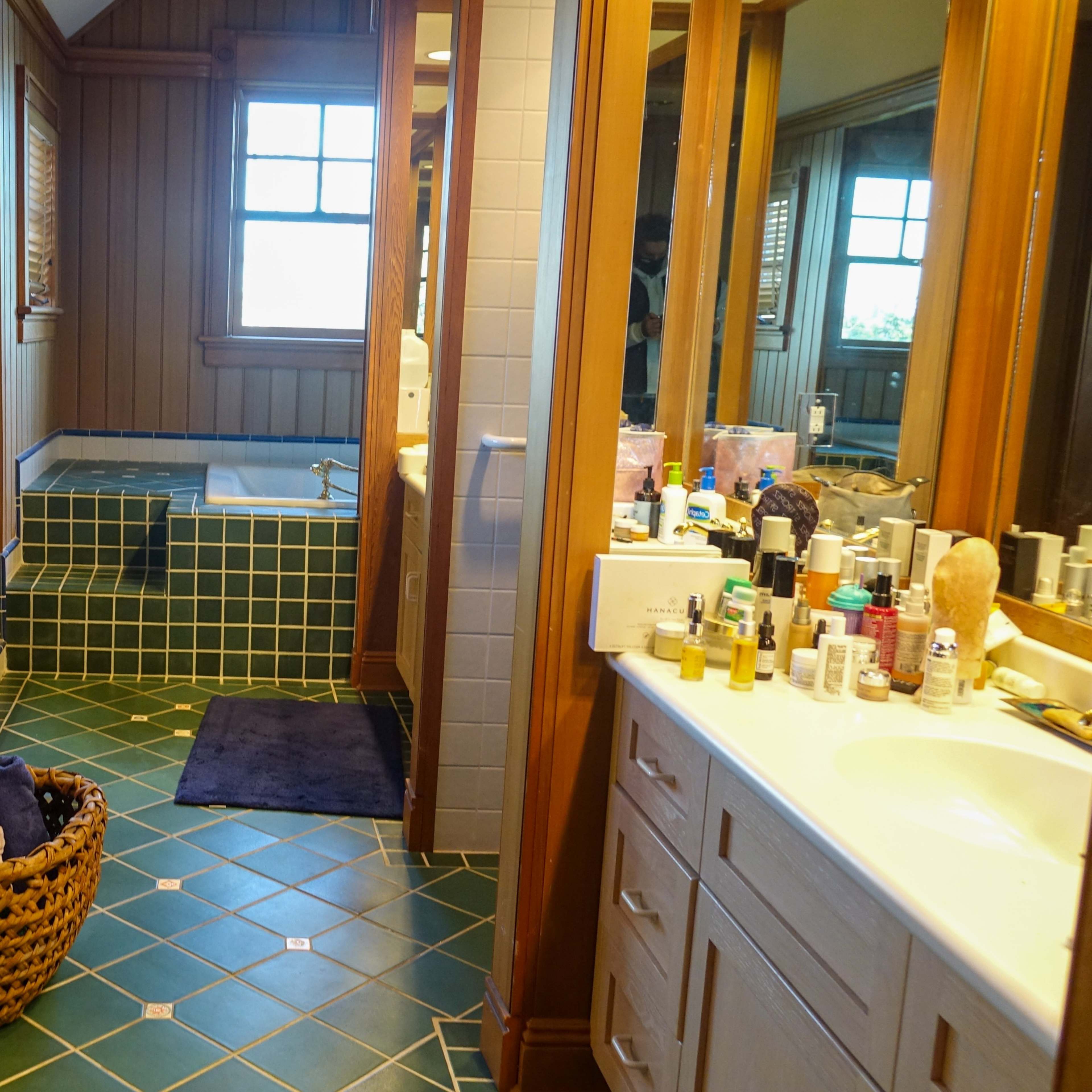 A spacious bathroom featuring a green tiled floor, a wooden cabinet, a large mirror, and two sinks with various toiletries displayed.