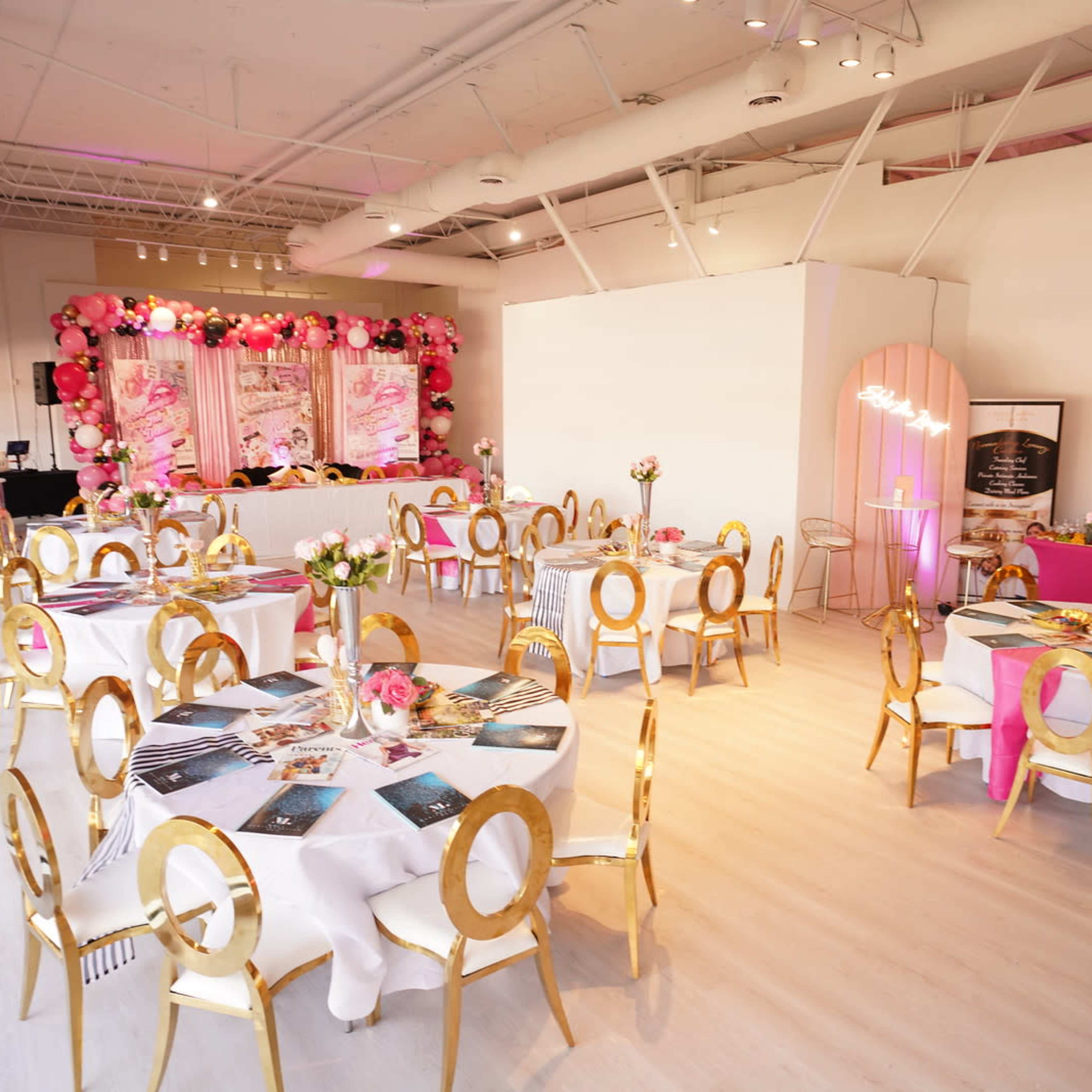 A well-decorated event space with round tables set for guests, featuring gold chairs and floral centerpieces, and a backdrop with pink decorations.