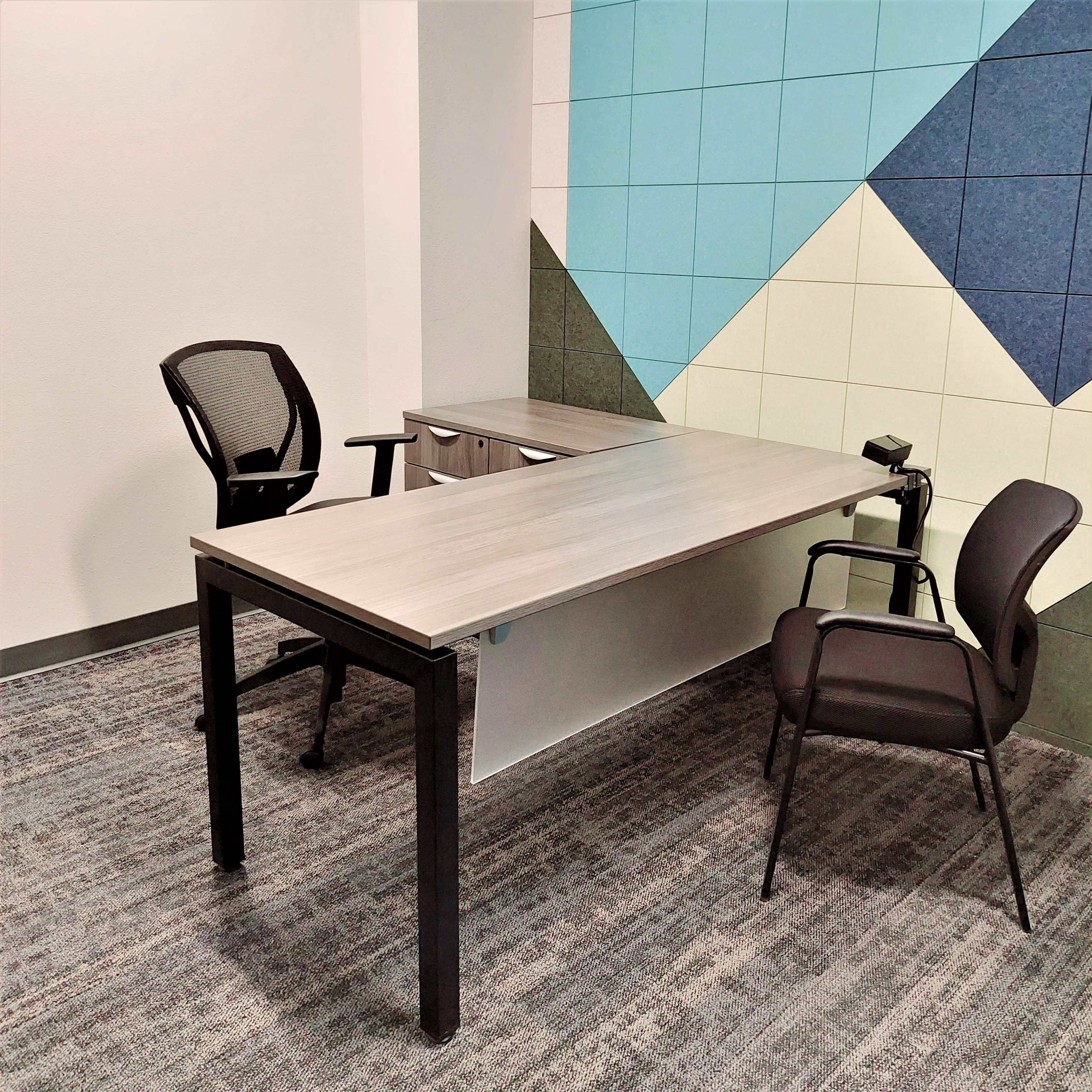 A sparsely furnished office space featuring a large table and two office chairs against a colorful wall of geometric shapes.
