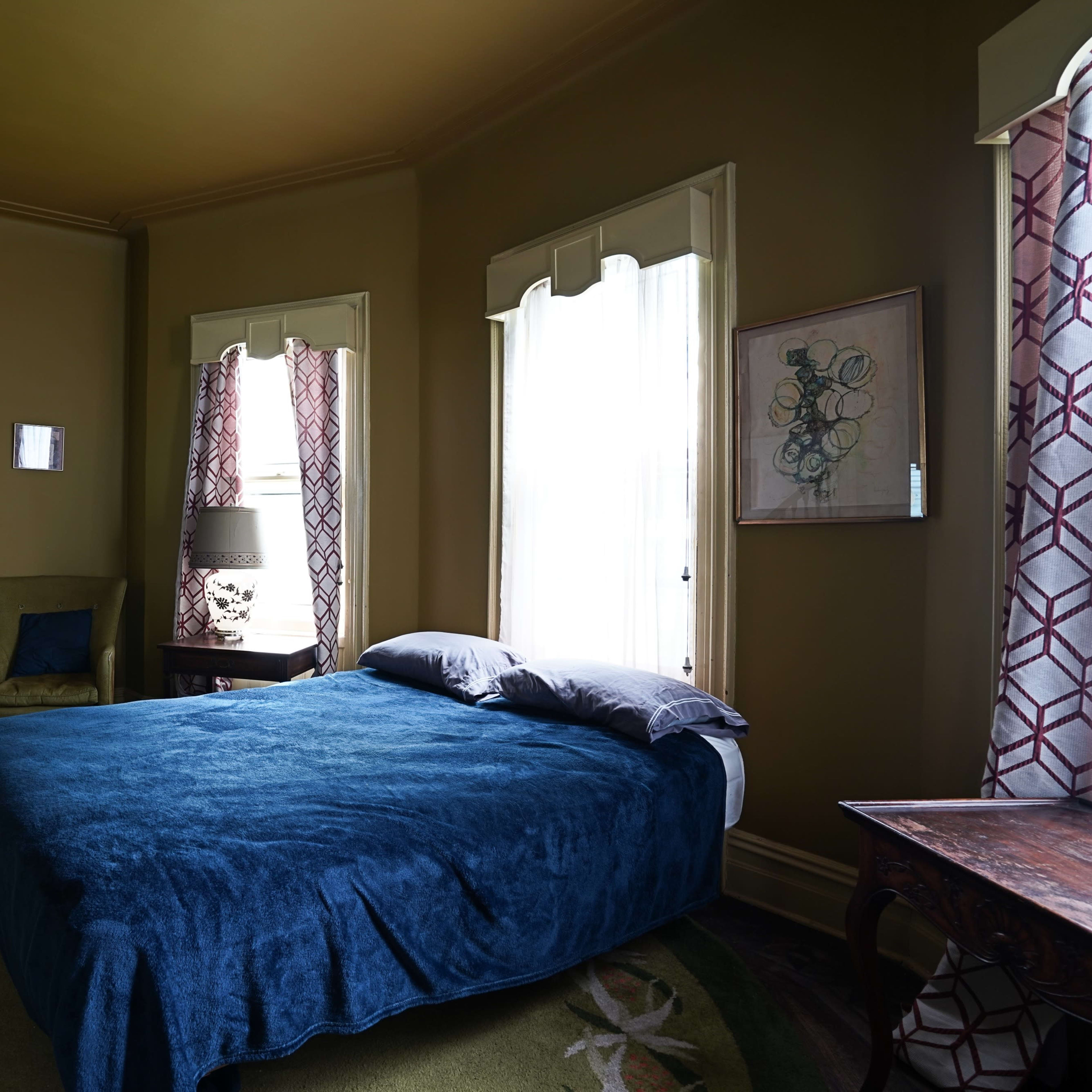 A bedroom features a bed with a blue blanket, accompanied by two windows and a view into a living area with a sofa.