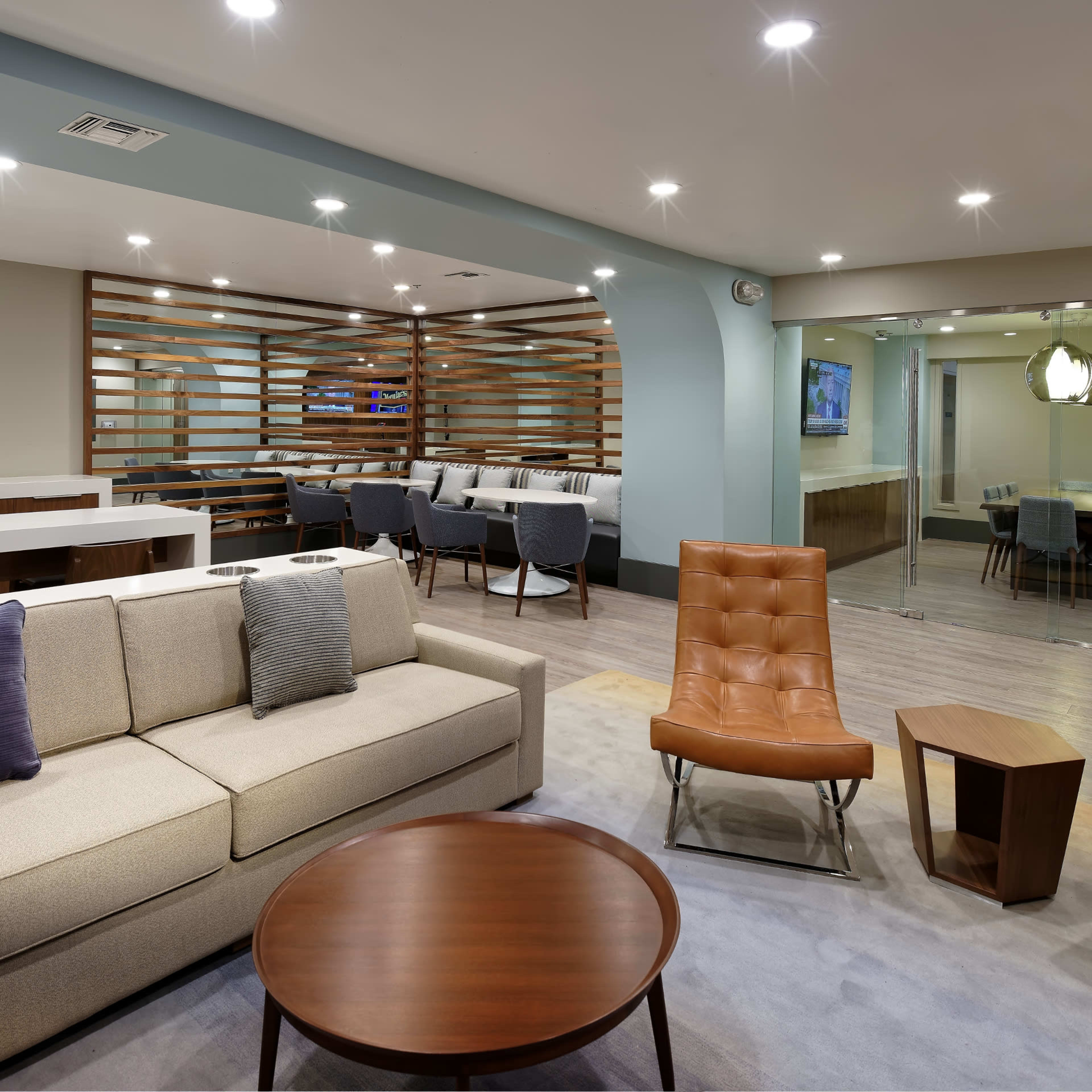 A modern lounge area featuring a beige couch, two orange chairs, a round wooden coffee table, and a dining area with wooden partitioning in the background.