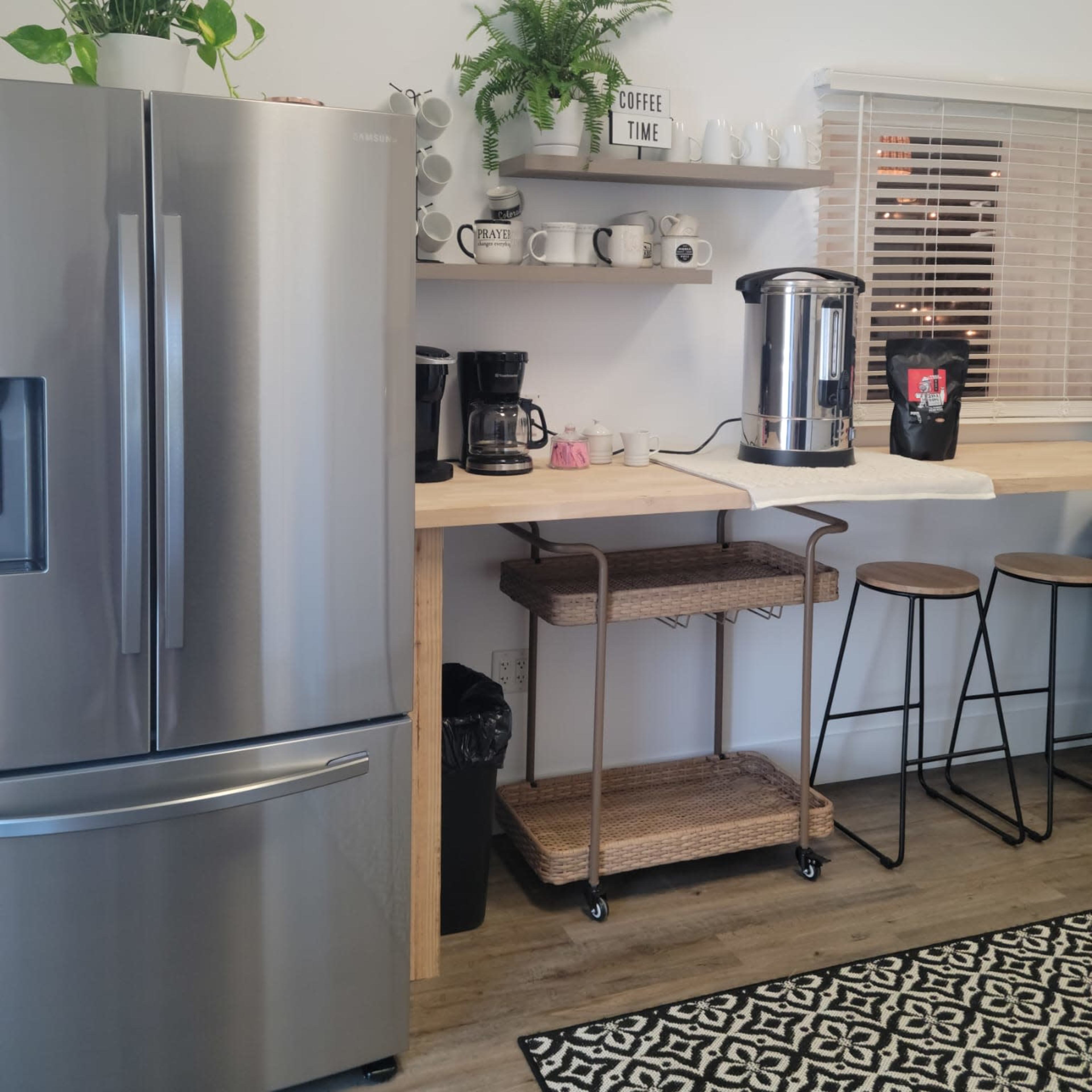 A modern kitchen features a stainless steel refrigerator, a coffee station with multiple coffee makers, and a bar with stools, all arranged against a bright, minimalistic backdrop.