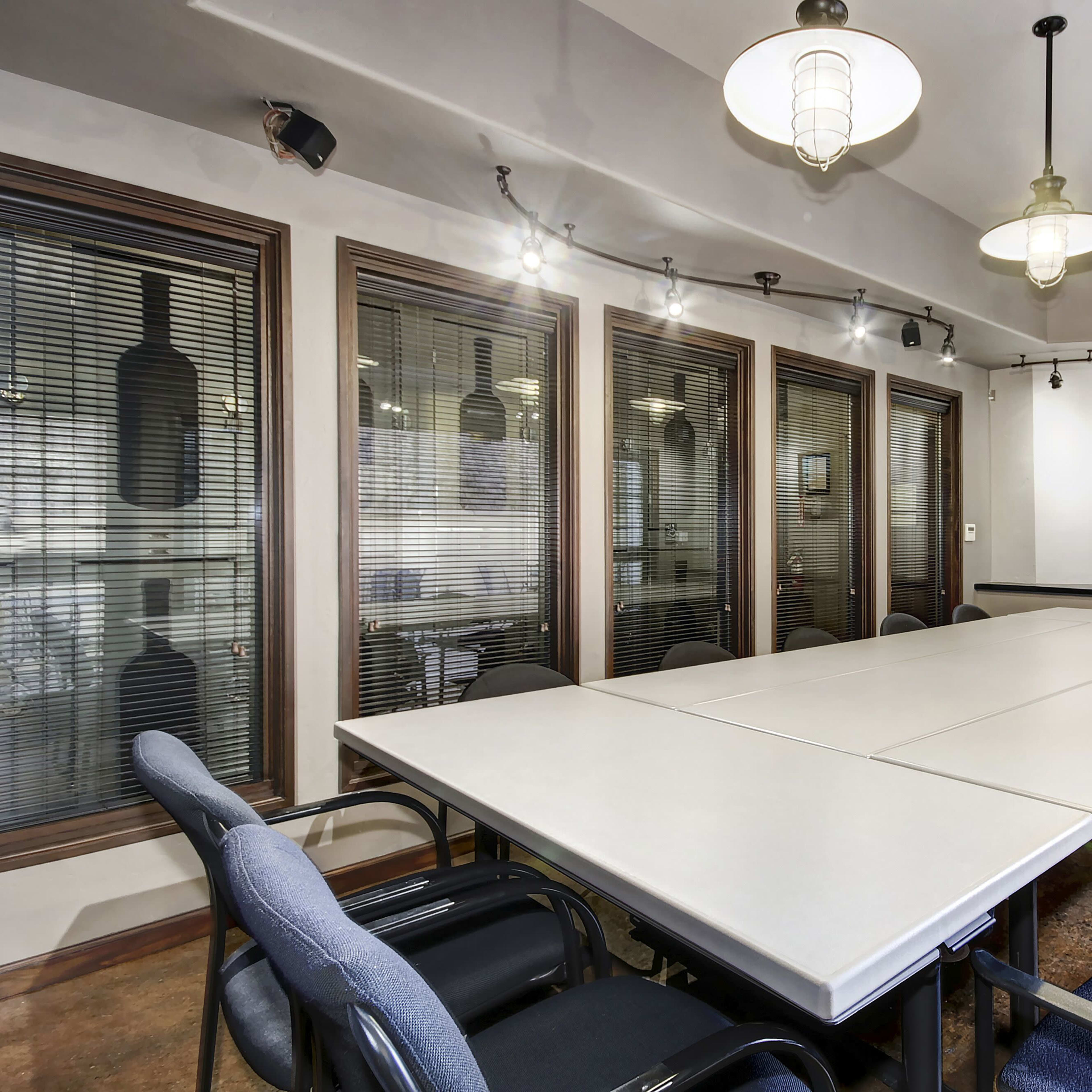A conference room features a large rectangular table surrounded by black and blue chairs, with windows on one side and pendant lighting above.
