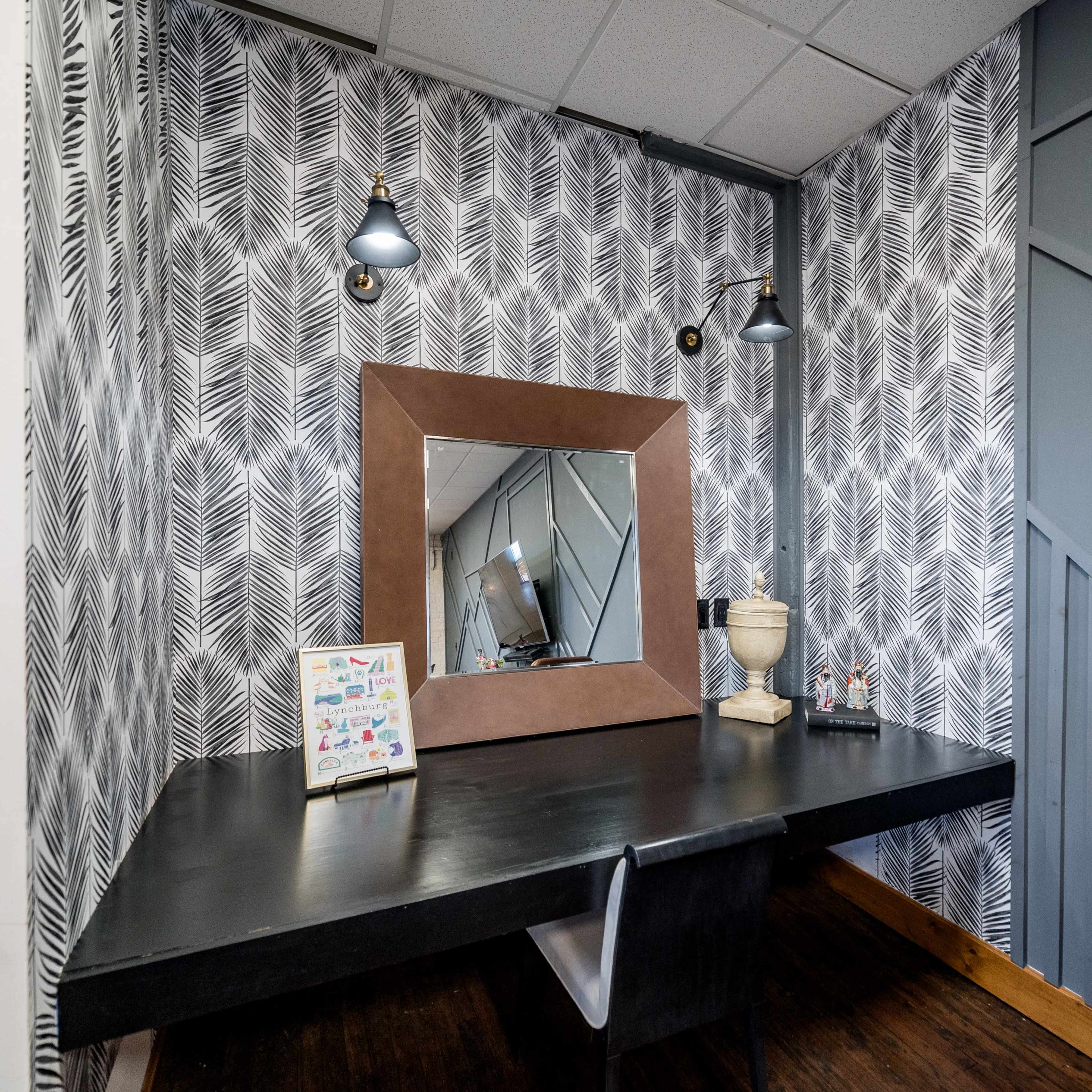 A modern desk with a large mirror is positioned against a wallpapered wall featuring a leaf pattern, illuminated by two wall sconces.