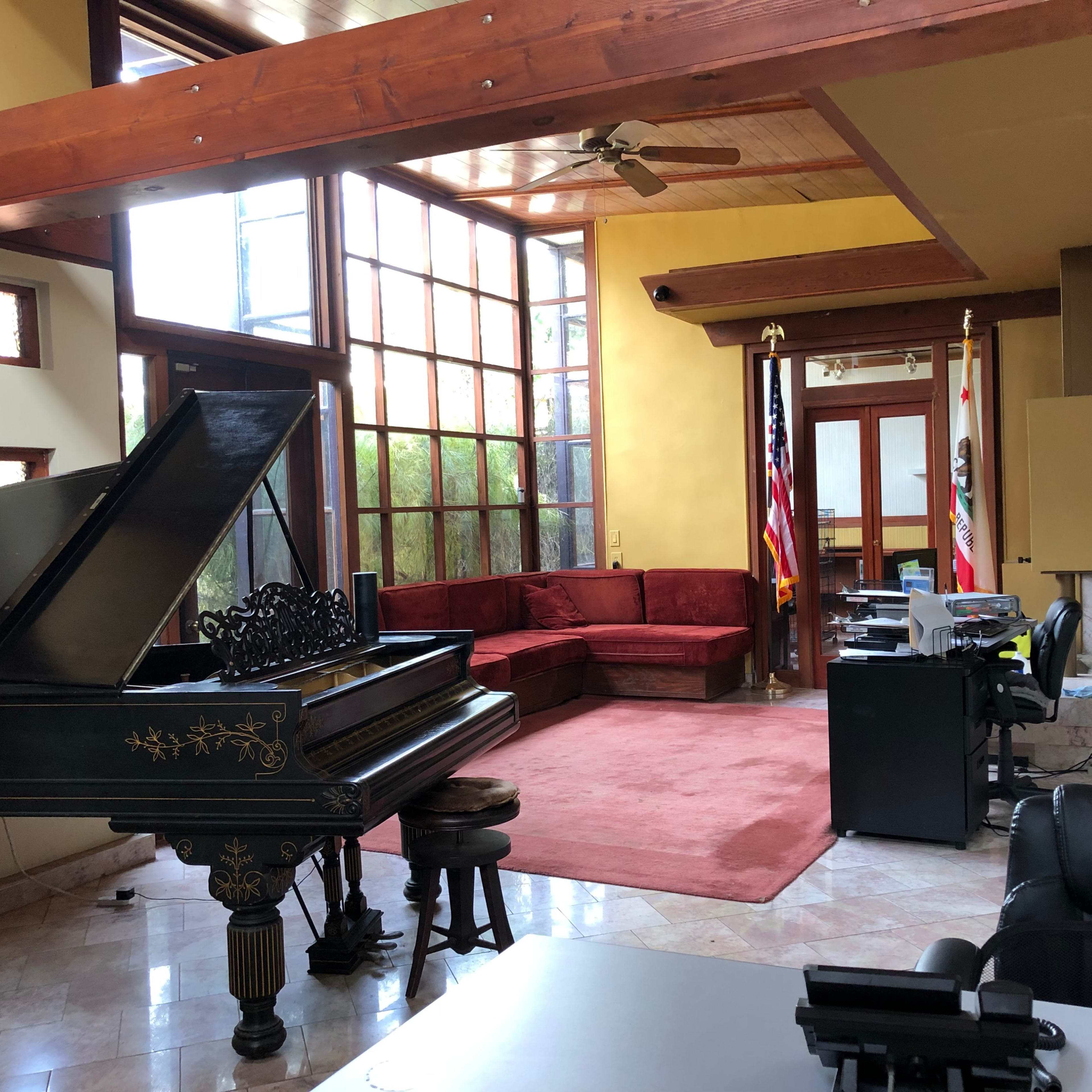 A spacious room featuring a grand piano, a red sectional sofa, a desk with a computer, and large windows allowing natural light to enter.