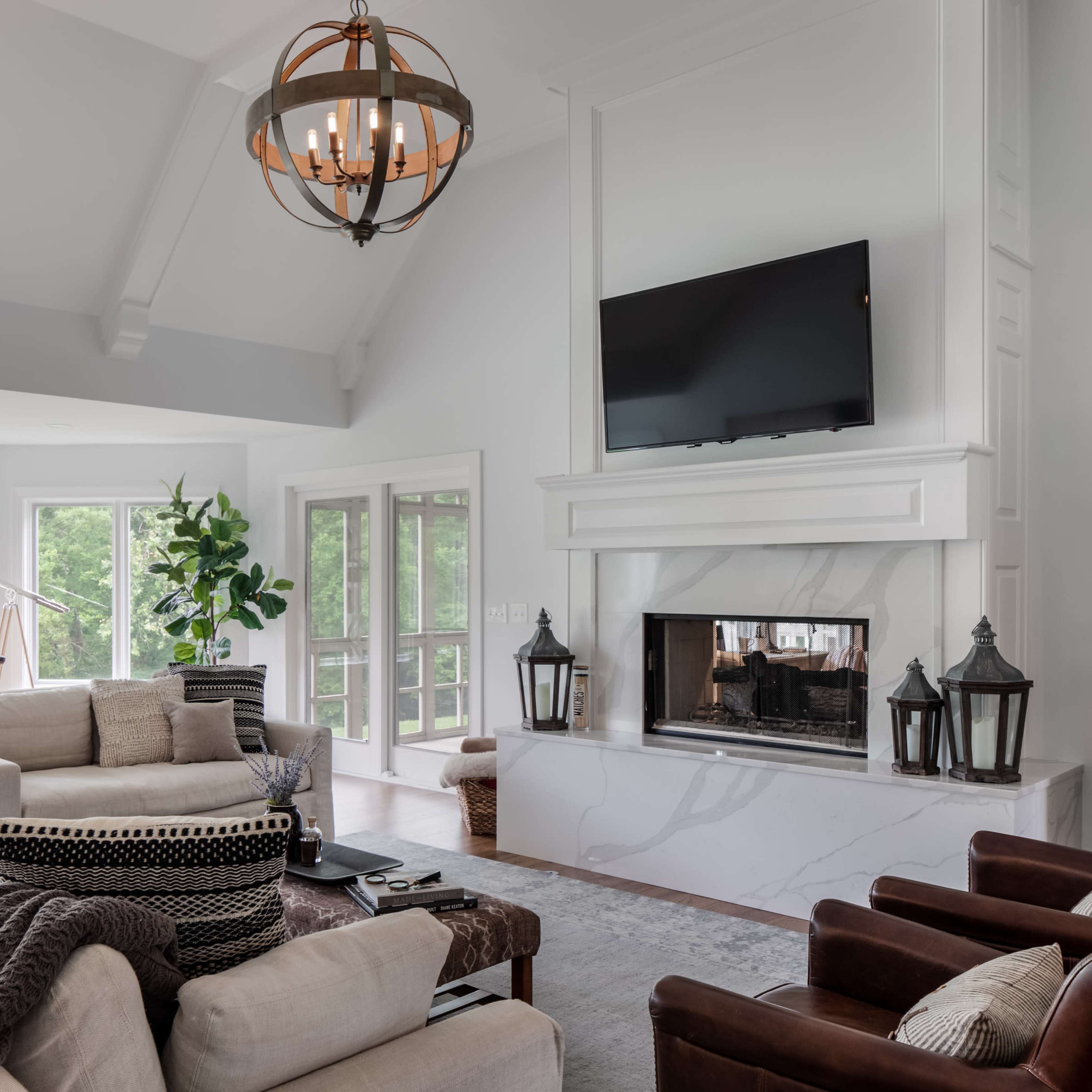A modern living room featuring a large sofa, armchairs, a fireplace with a television above it, and large windows that provide natural light.