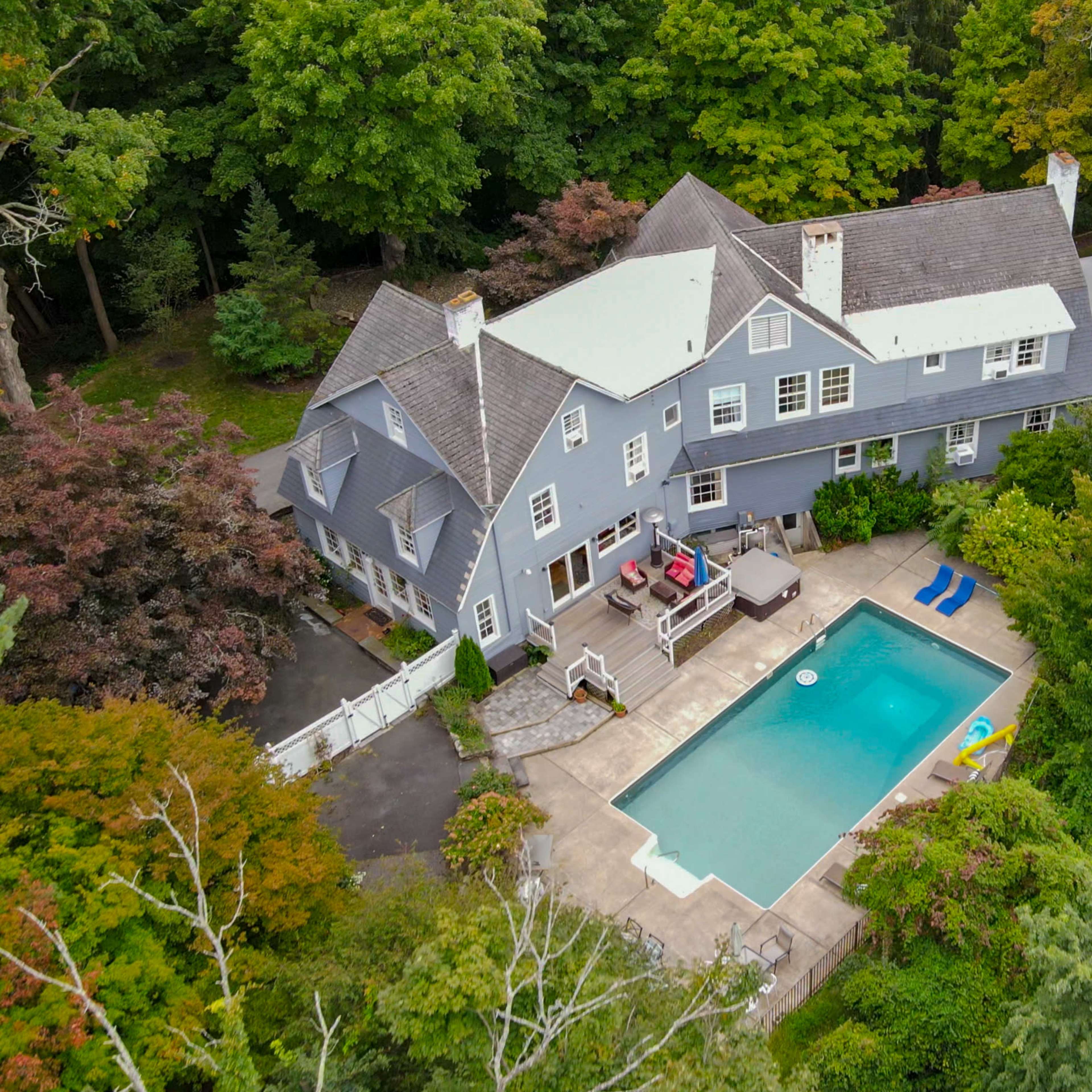 A large blue residential house with an adjacent rectangular swimming pool surrounded by a patio and lush greenery.