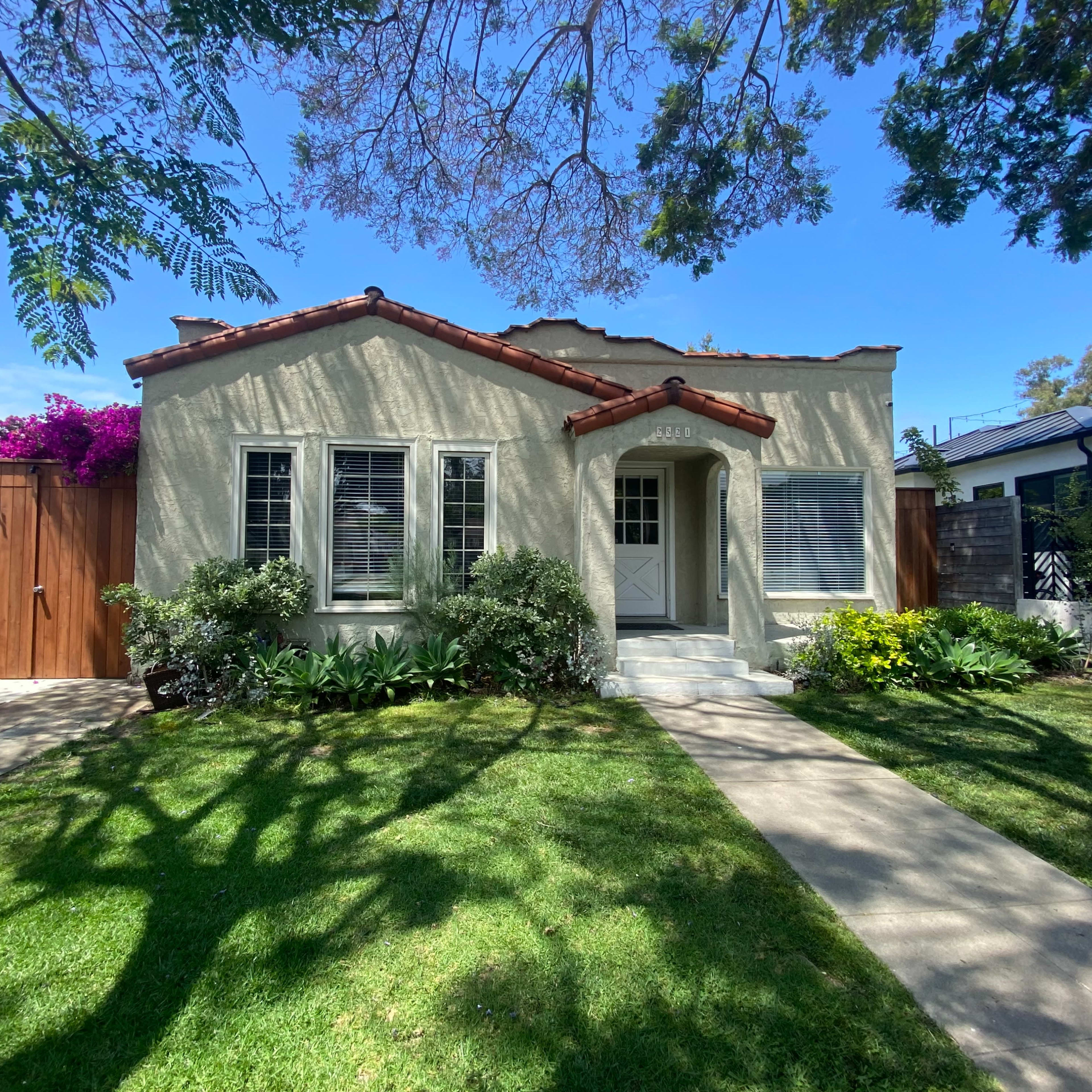 A single-story house with a light-colored exterior, a red-tiled roof, and a well-maintained front yard featuring shrubs and flowering plants.