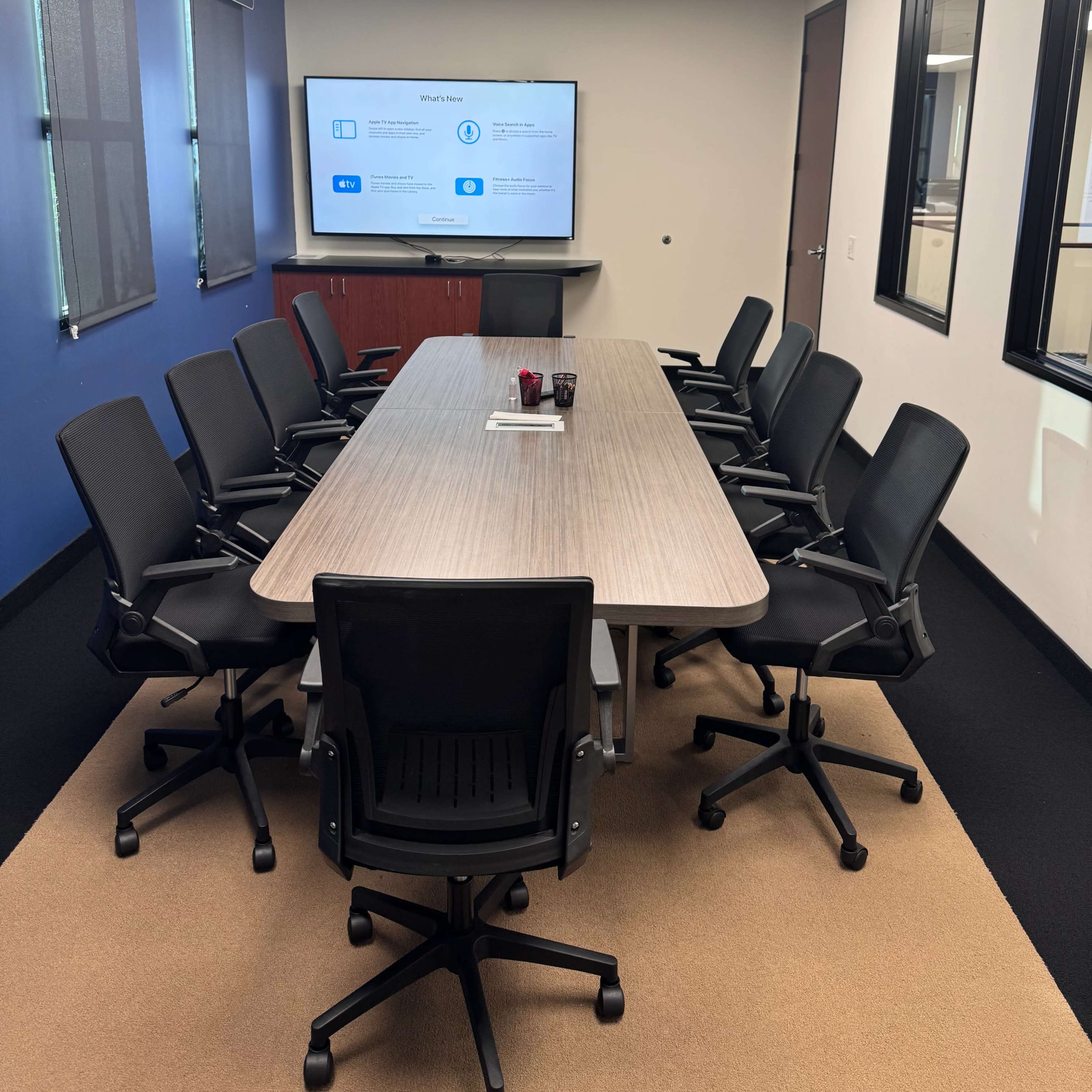 A modern conference room features a long table surrounded by black chairs, with a large screen displaying a presentation on the wall.