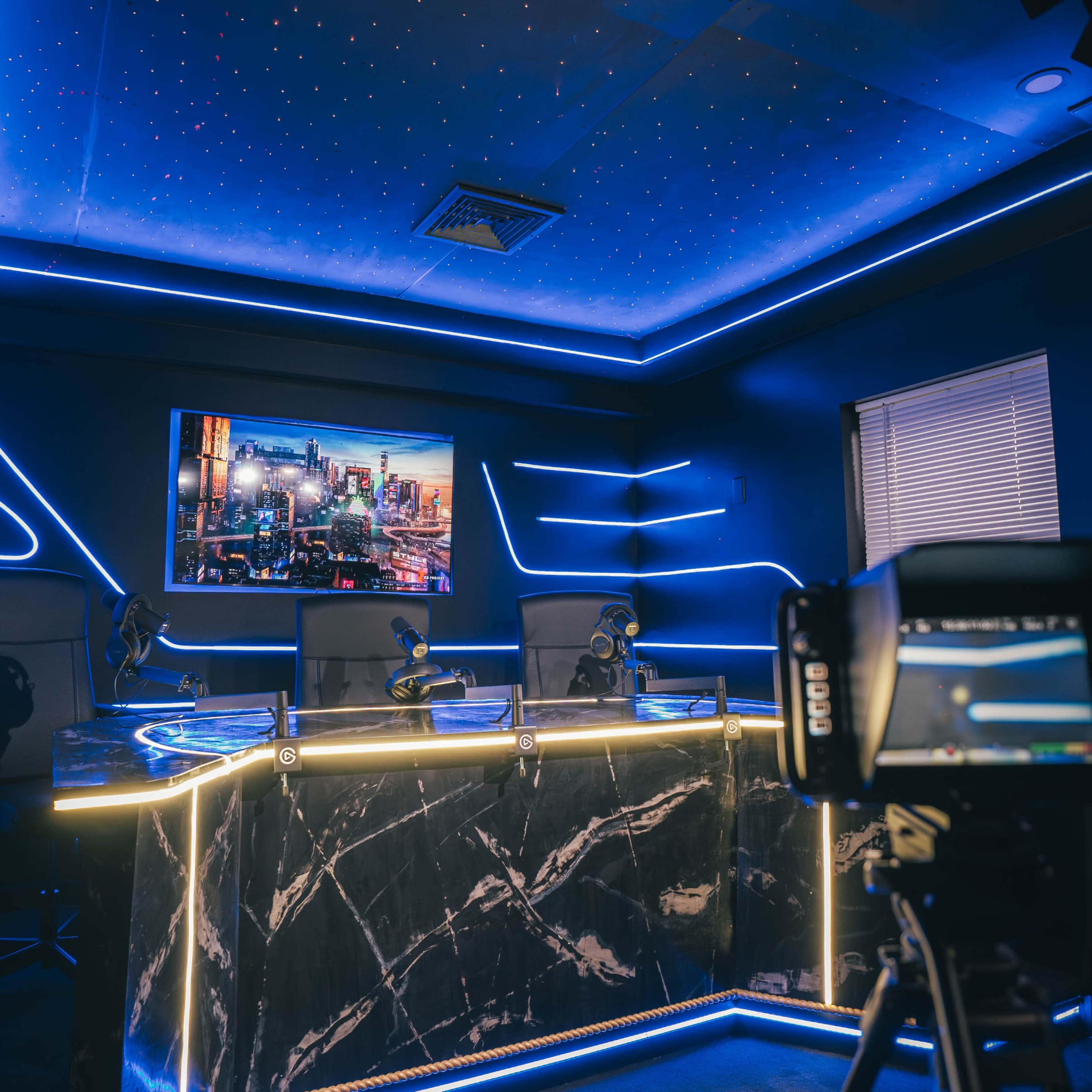 A modern studio setup featuring a sleek desk with microphones, a large screen displaying a cityscape, and ambient blue lighting.
