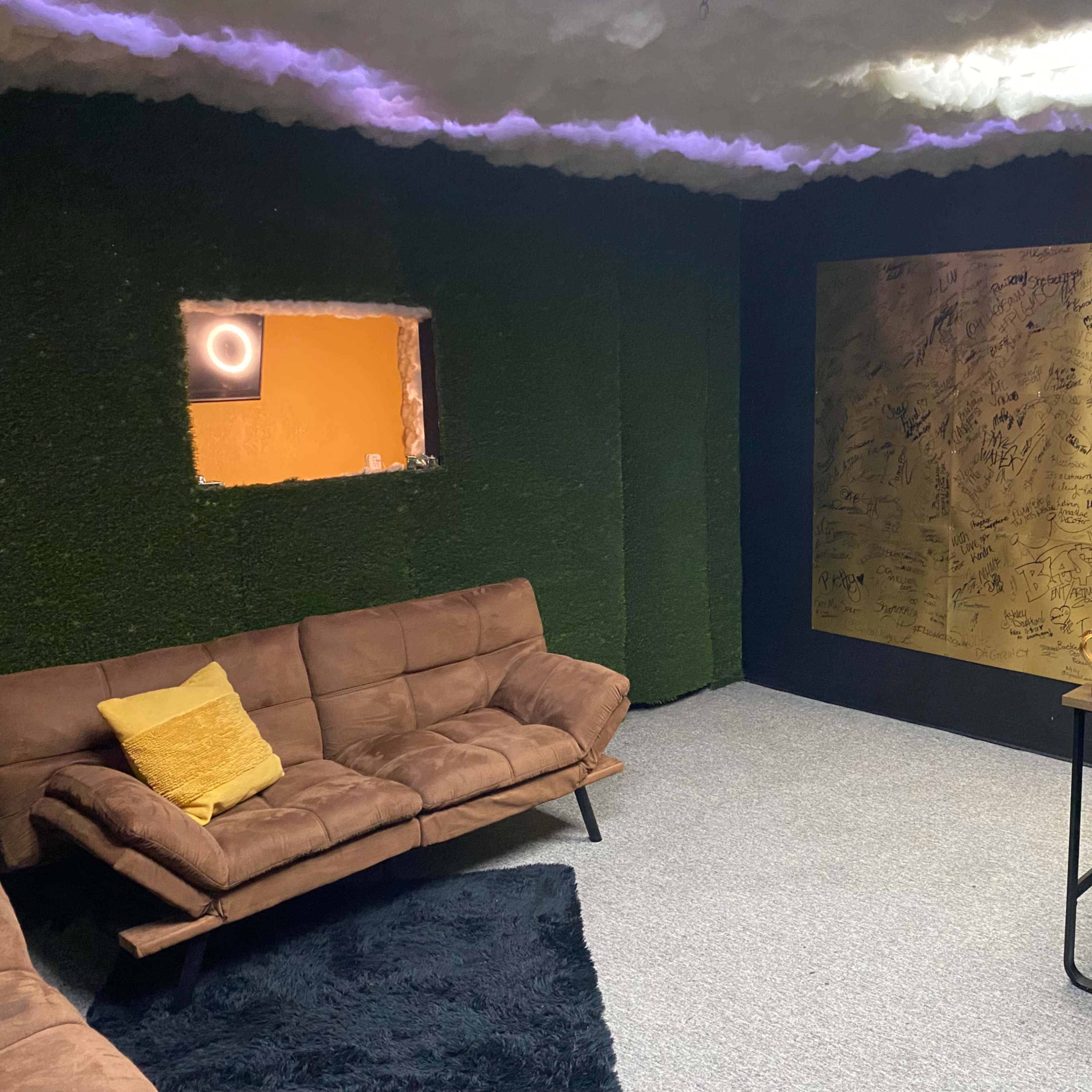 A modern room with a green moss-covered wall, brown couches, a gold wall covered in signatures, and a desk with a microphone setup.