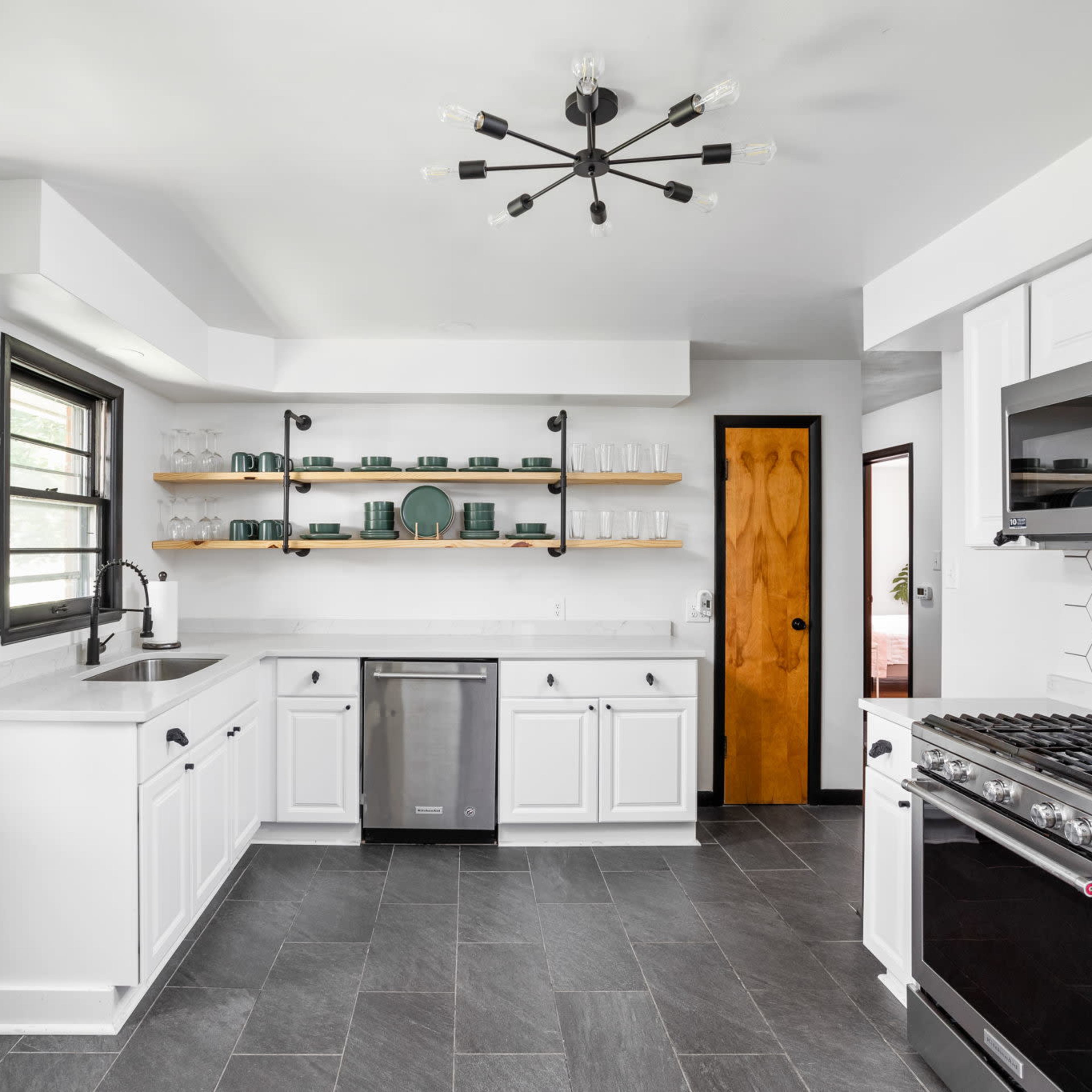 A modern kitchen features white cabinetry, a gray tile floor, open shelving with dishware, and a central light fixture.