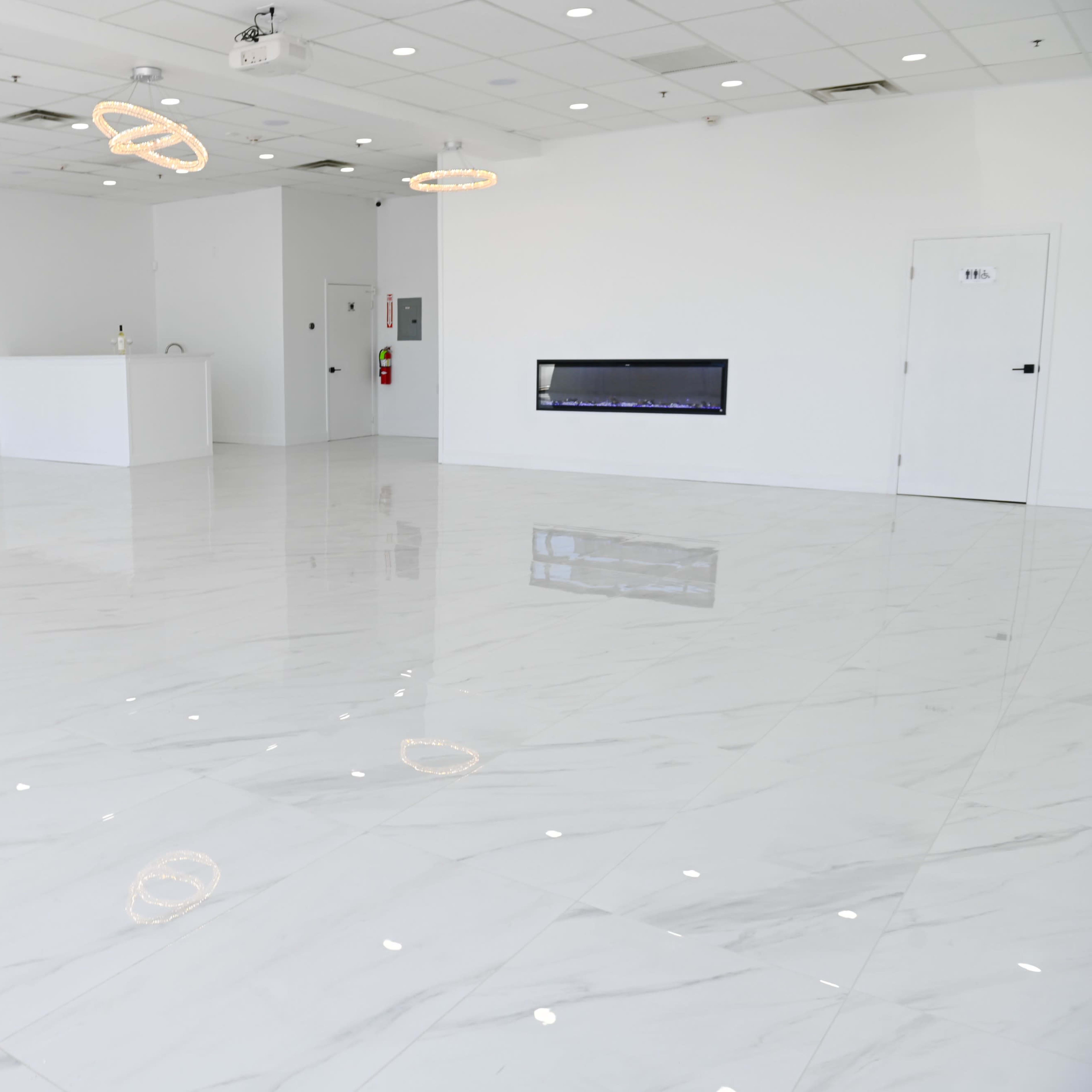 A spacious, empty room with white marble tiles, a recessed fireplace, and two circular light fixtures hanging from the ceiling.
