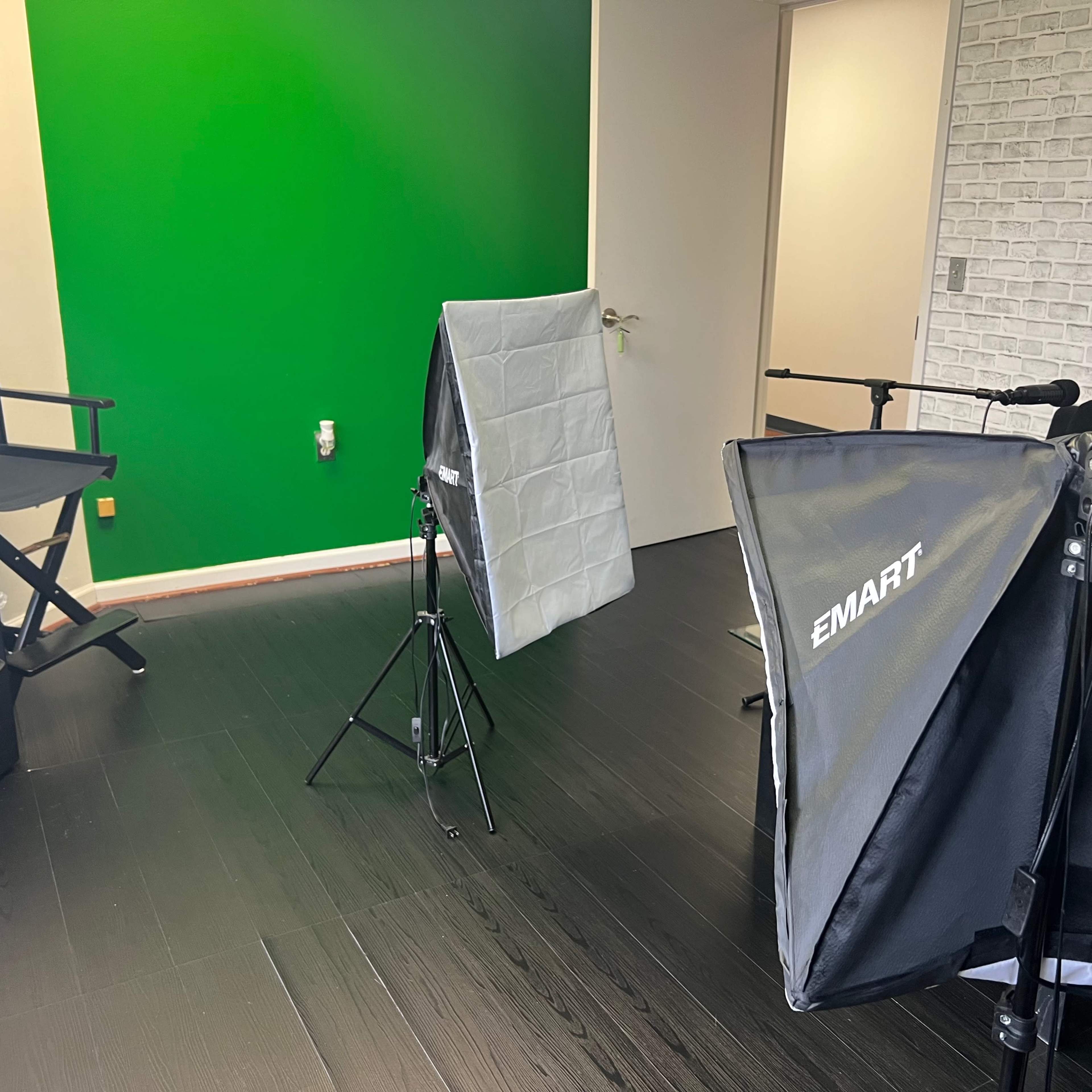 A studio setup with softbox lights, a green screen, and a chair in a room with dark flooring and a white brick wall.