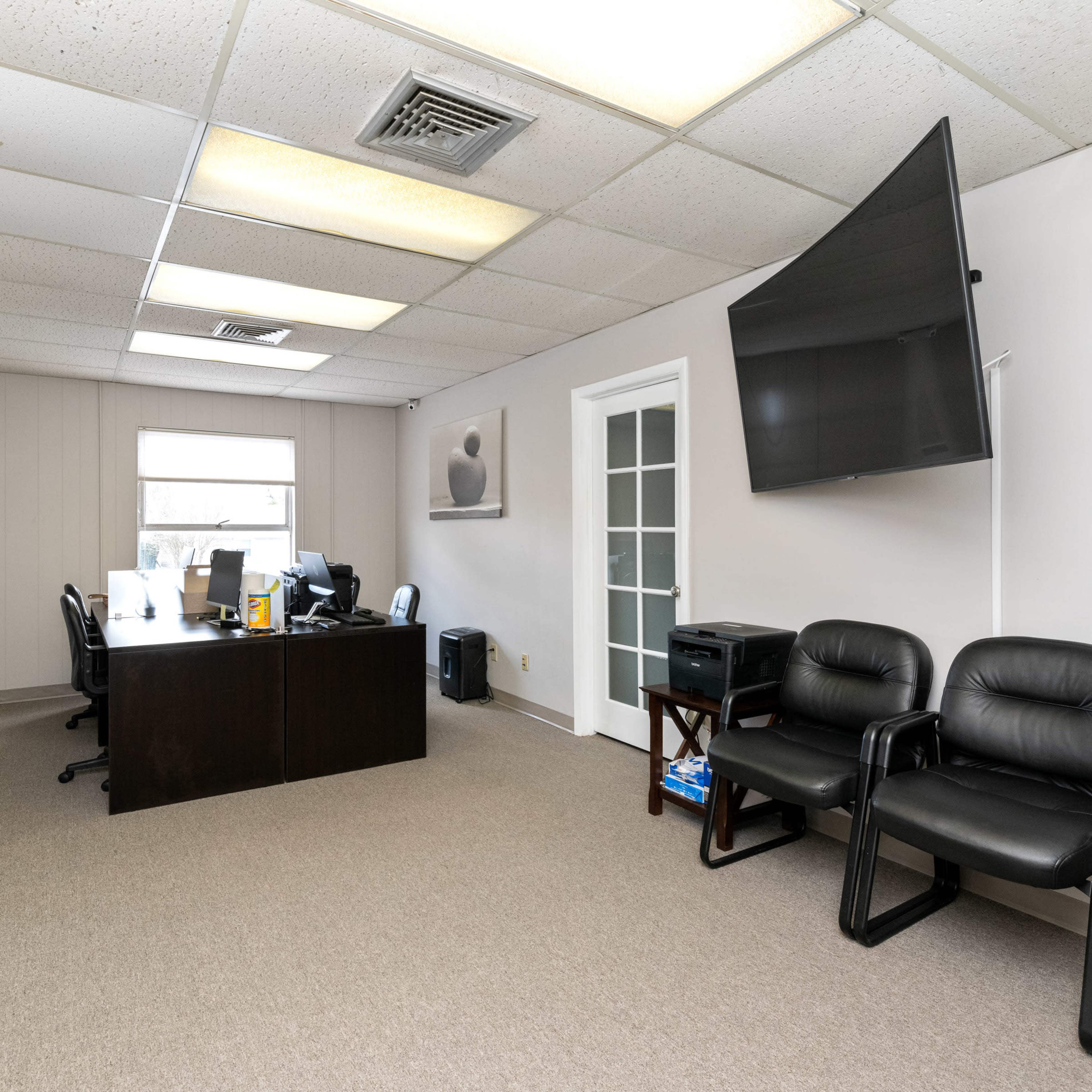 A spacious office with a large desk, an overhead TV, and two black chairs against the wall.