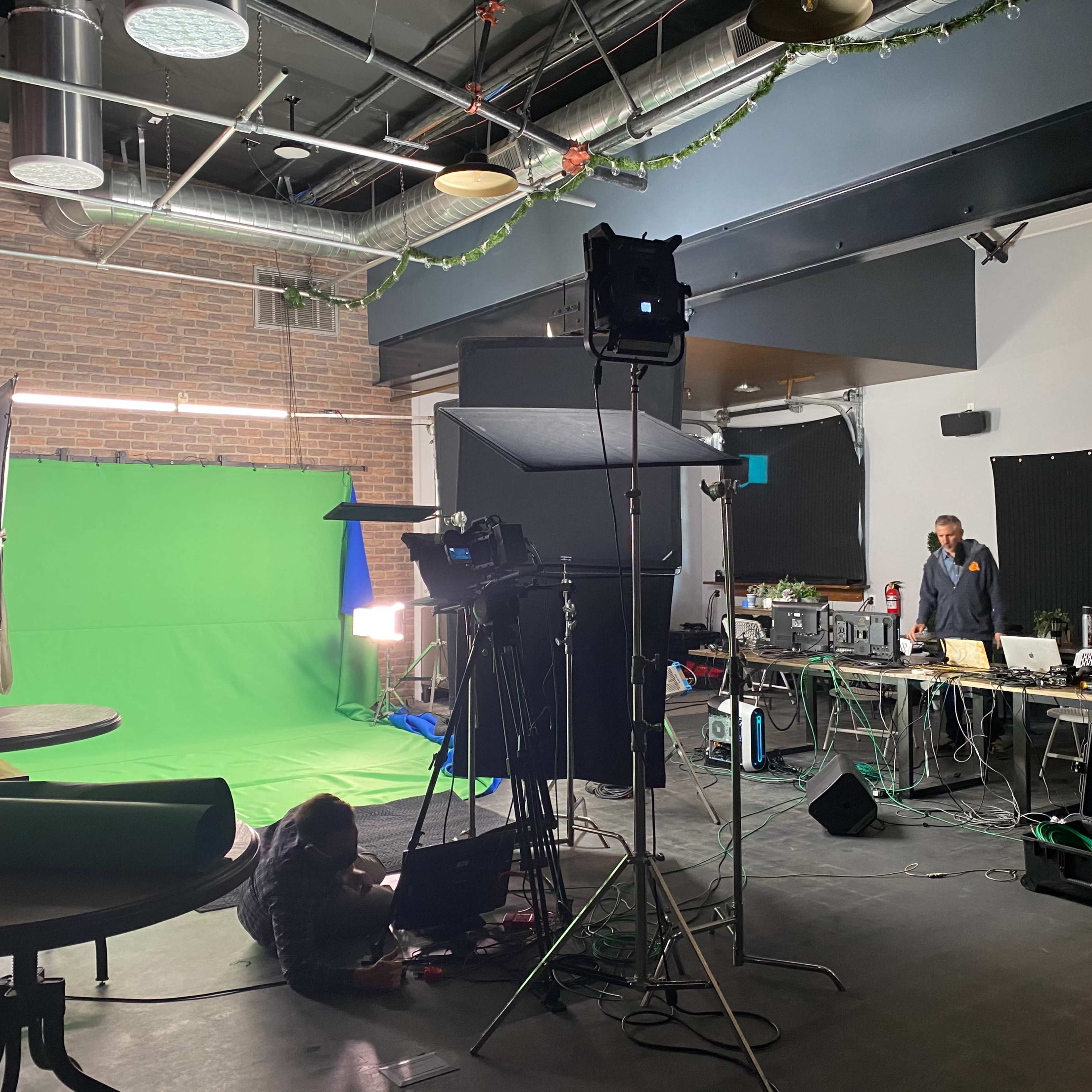 A studio space is set up with a green screen, lighting equipment, and several workstations, while a person is working on a laptop on the floor.