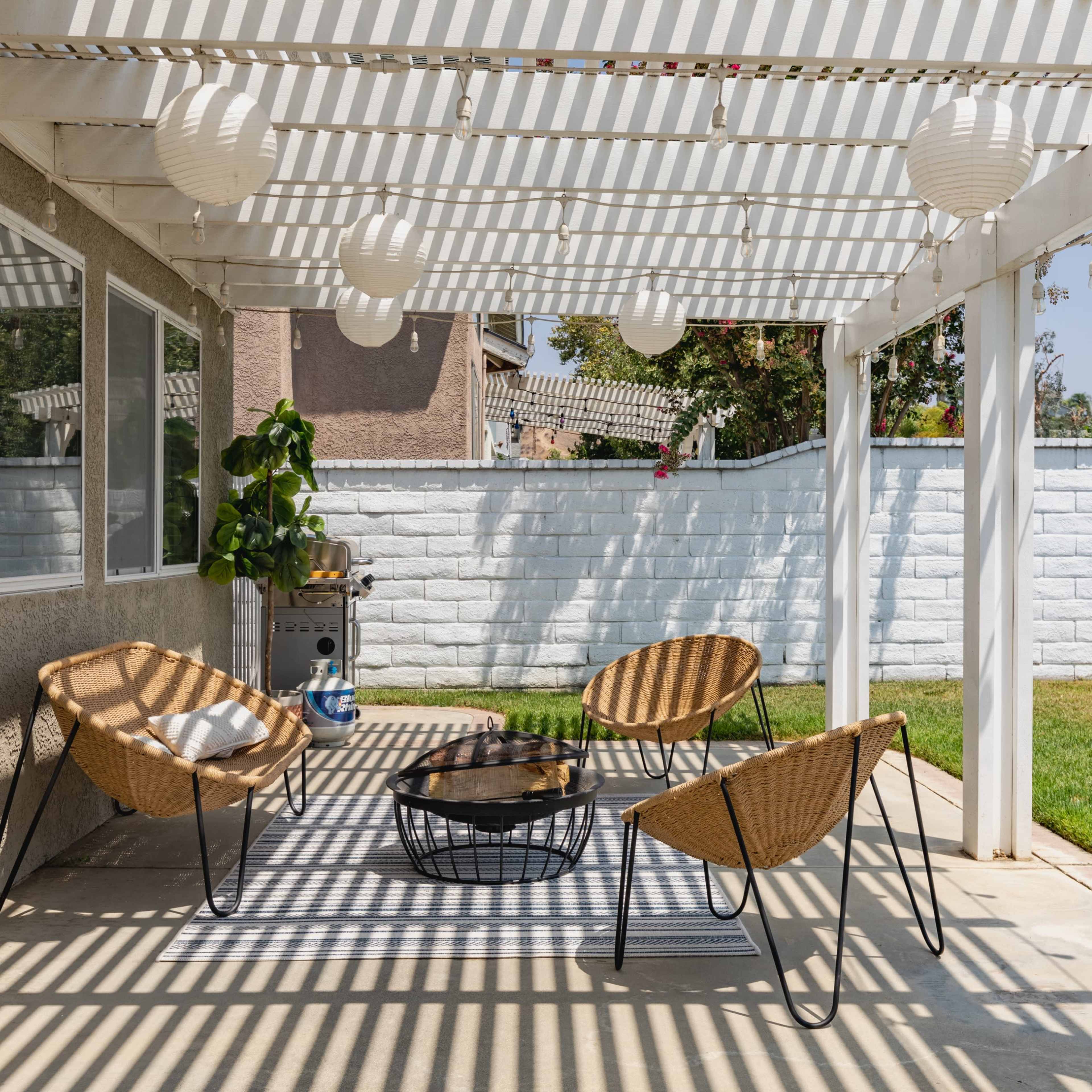 A patio features four woven chairs around a coffee table, with a striped rug underneath and lanterns hanging from a pergola.