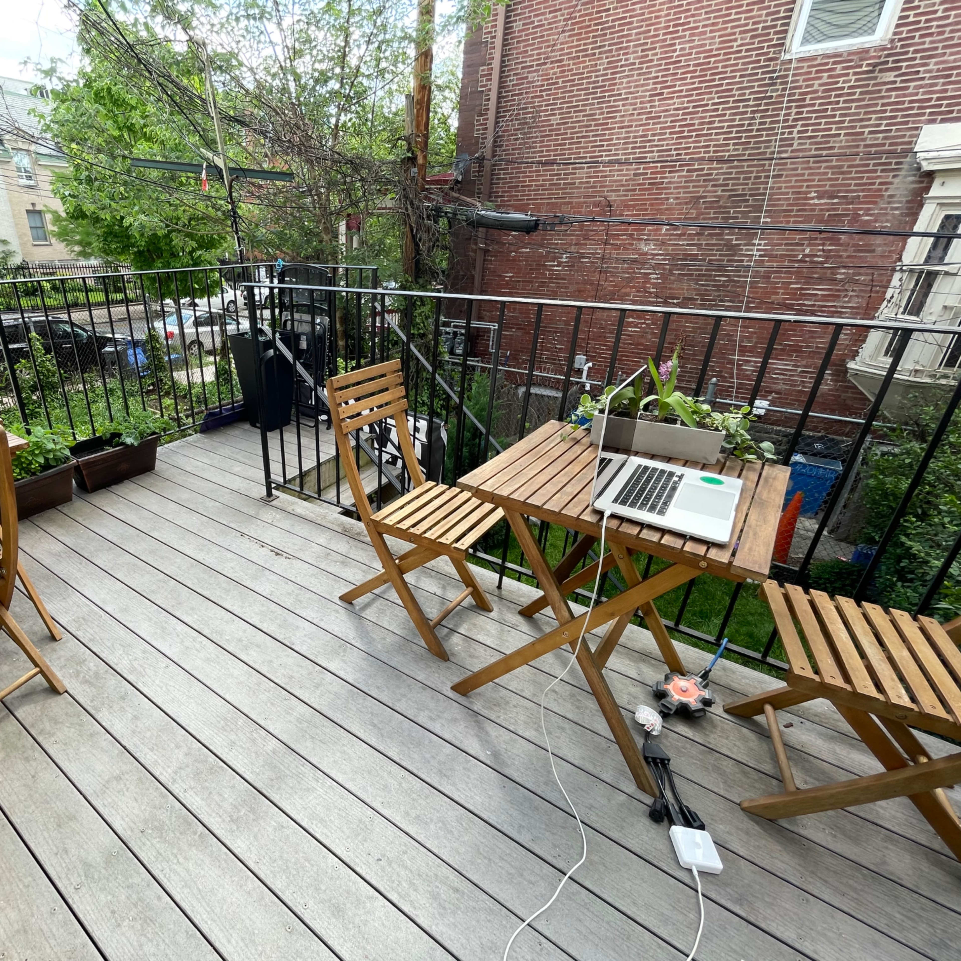 A small wooden deck with two folding tables and chairs, a laptop on one table, and potted plants along the edge.