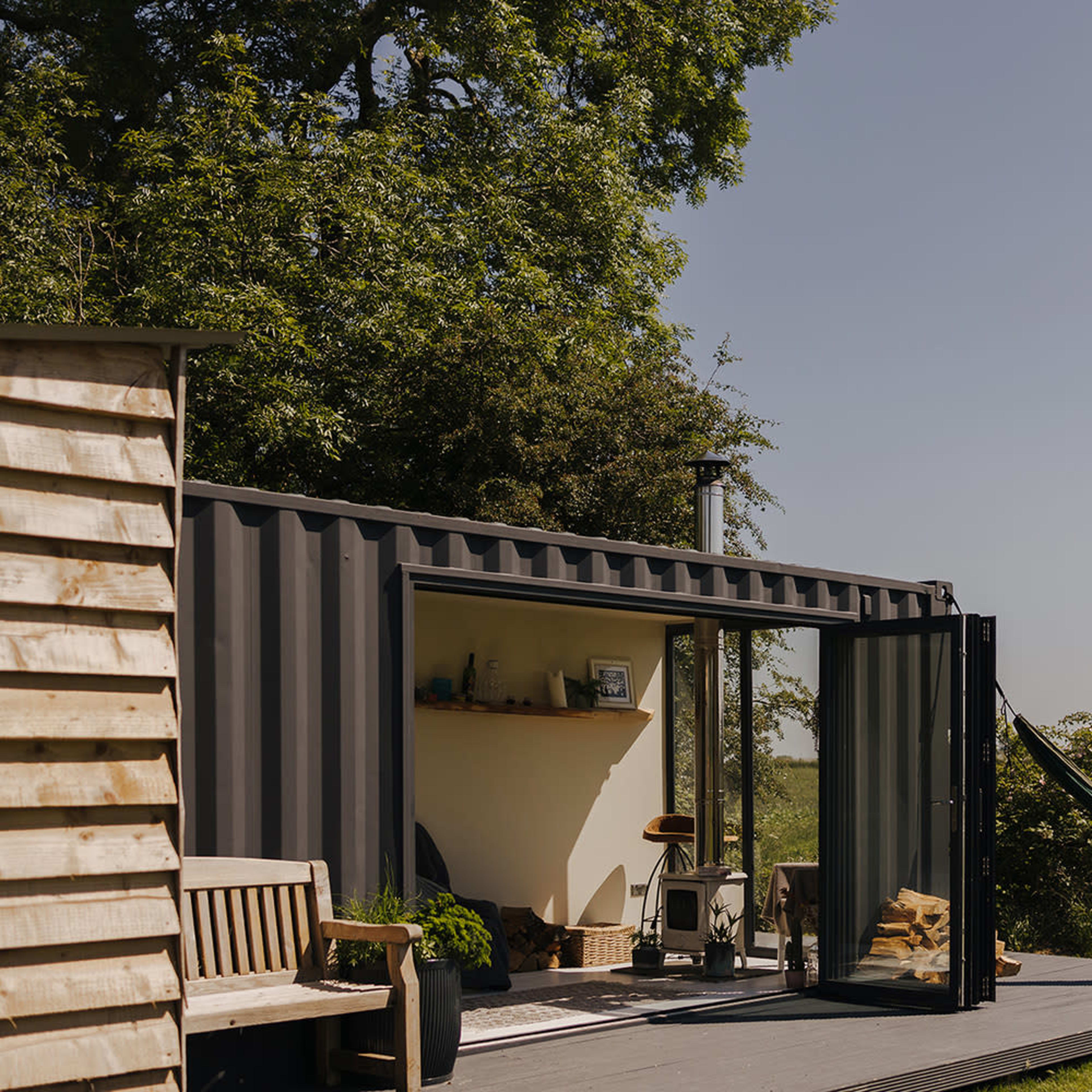 A modern, shipping container-style cabin sits on a deck with a hammock, surrounded by greenery and open fields.