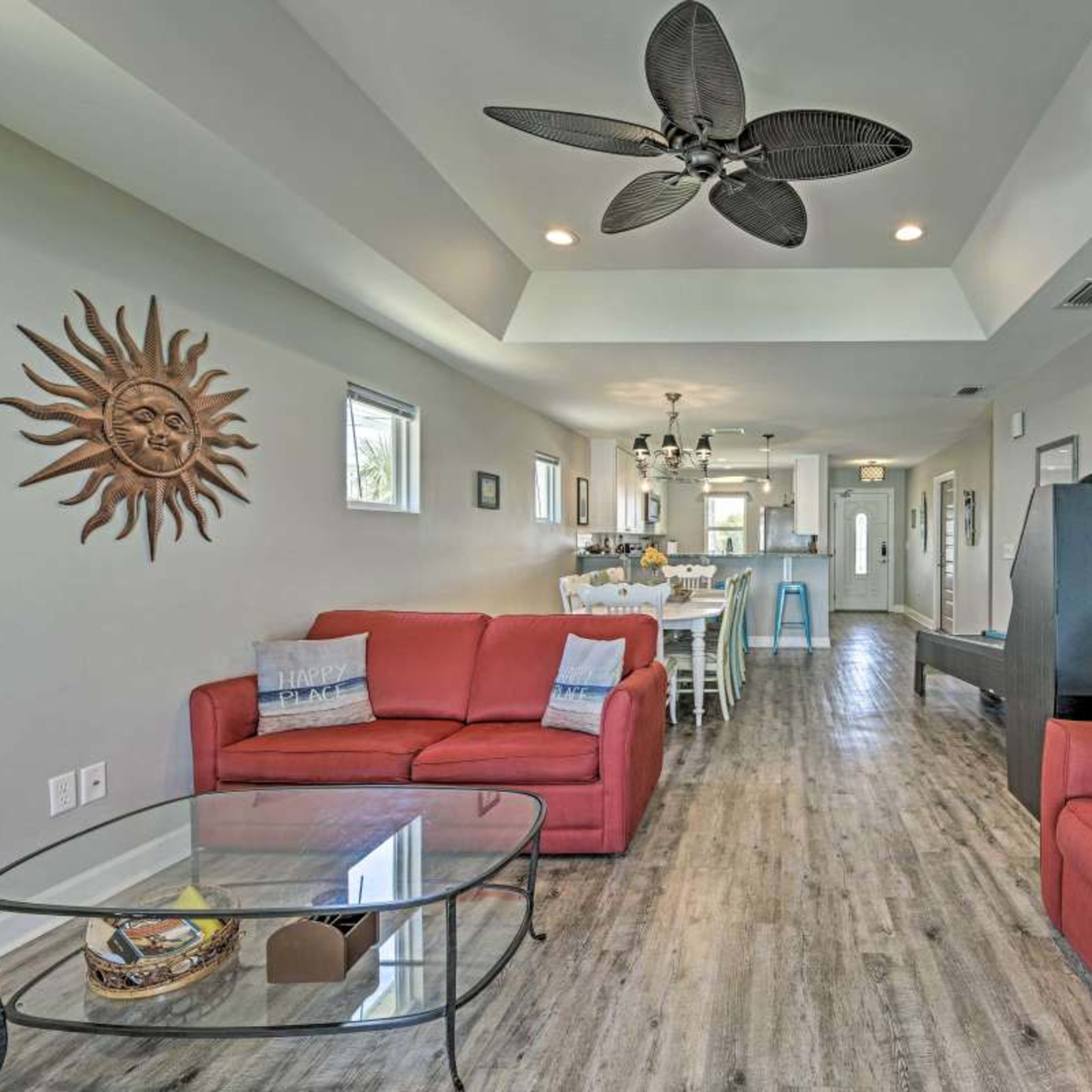 A living area with two red sofas, a glass coffee table, and a ceiling fan, leading into a dining space and a kitchen at the back.