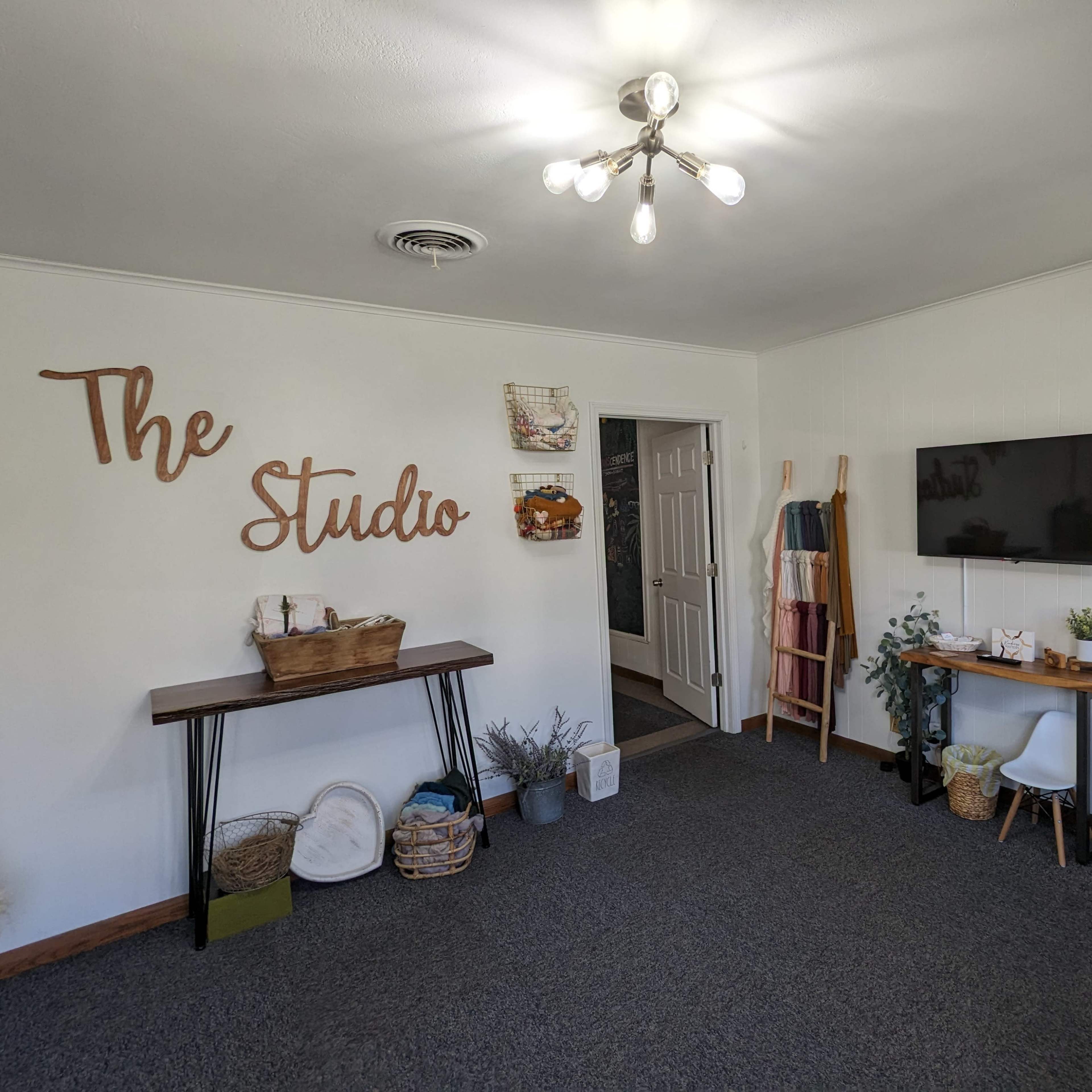 A room labeled "The Studio," featuring a wooden table, a wall-mounted television, and decorative items along with a gray stool.