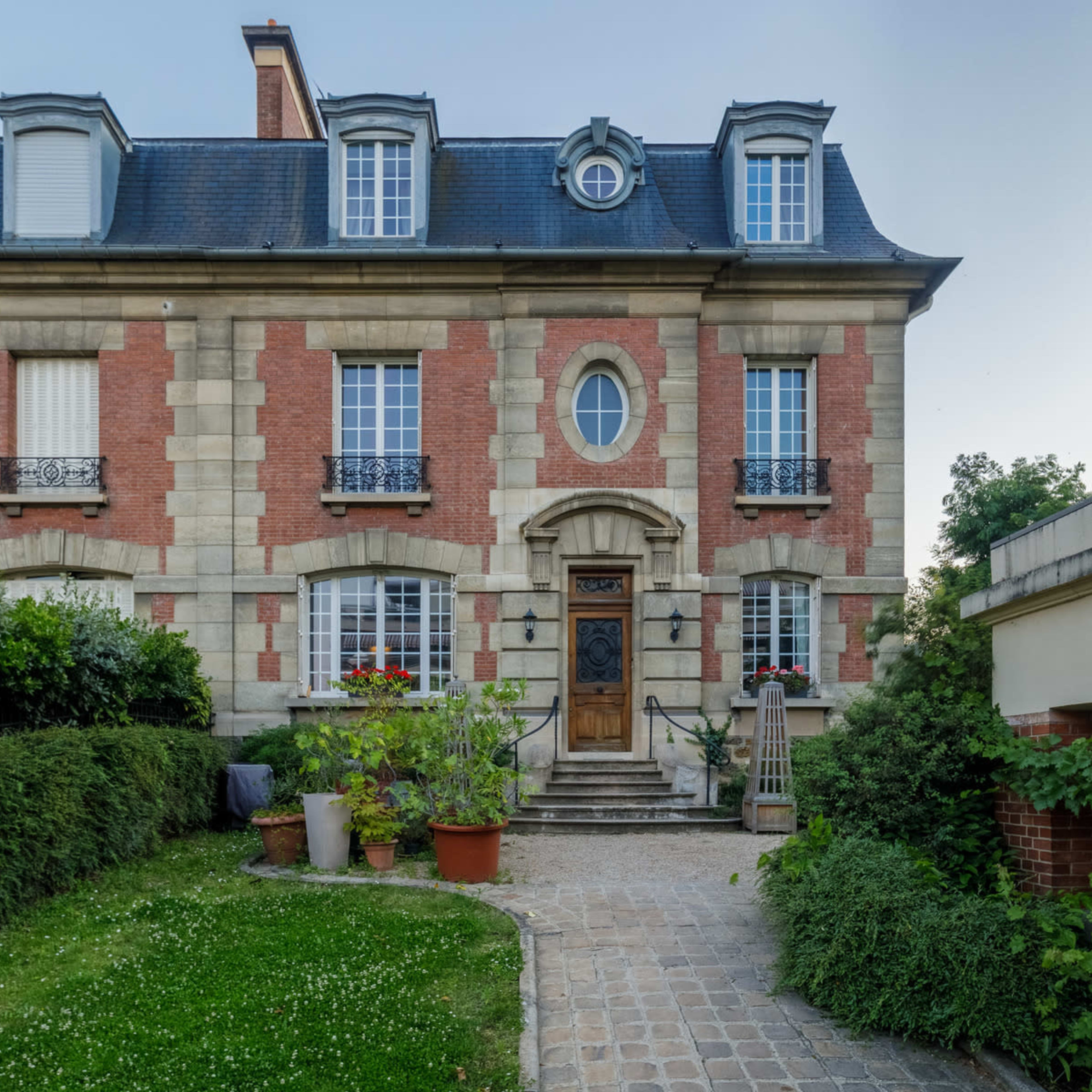 A three-story brick and stone house features a central wooden door, surrounded by garden beds and manicured hedges.
