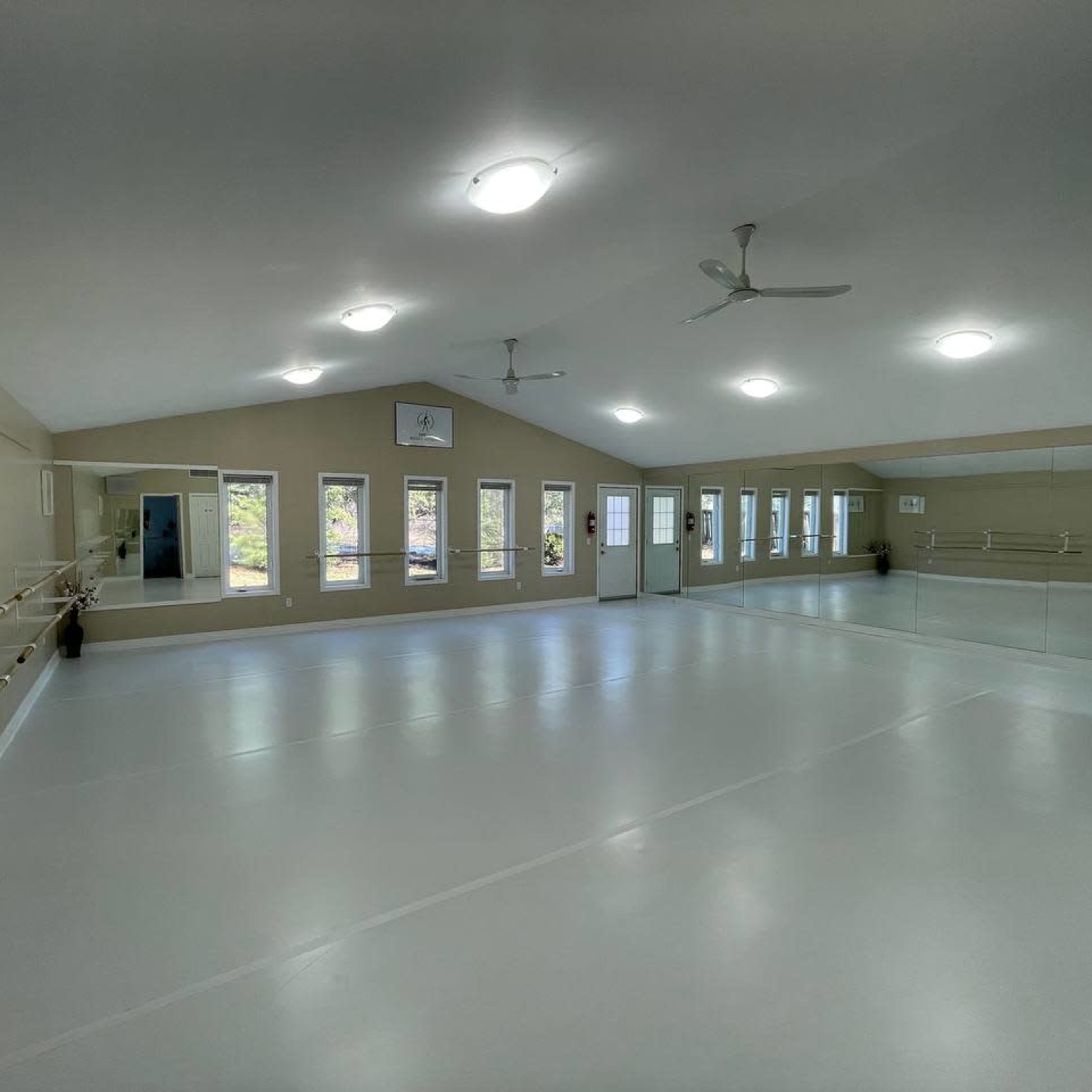 A bright dance studio features a smooth, white floor and large mirrors along one wall, with windows allowing natural light to enter.