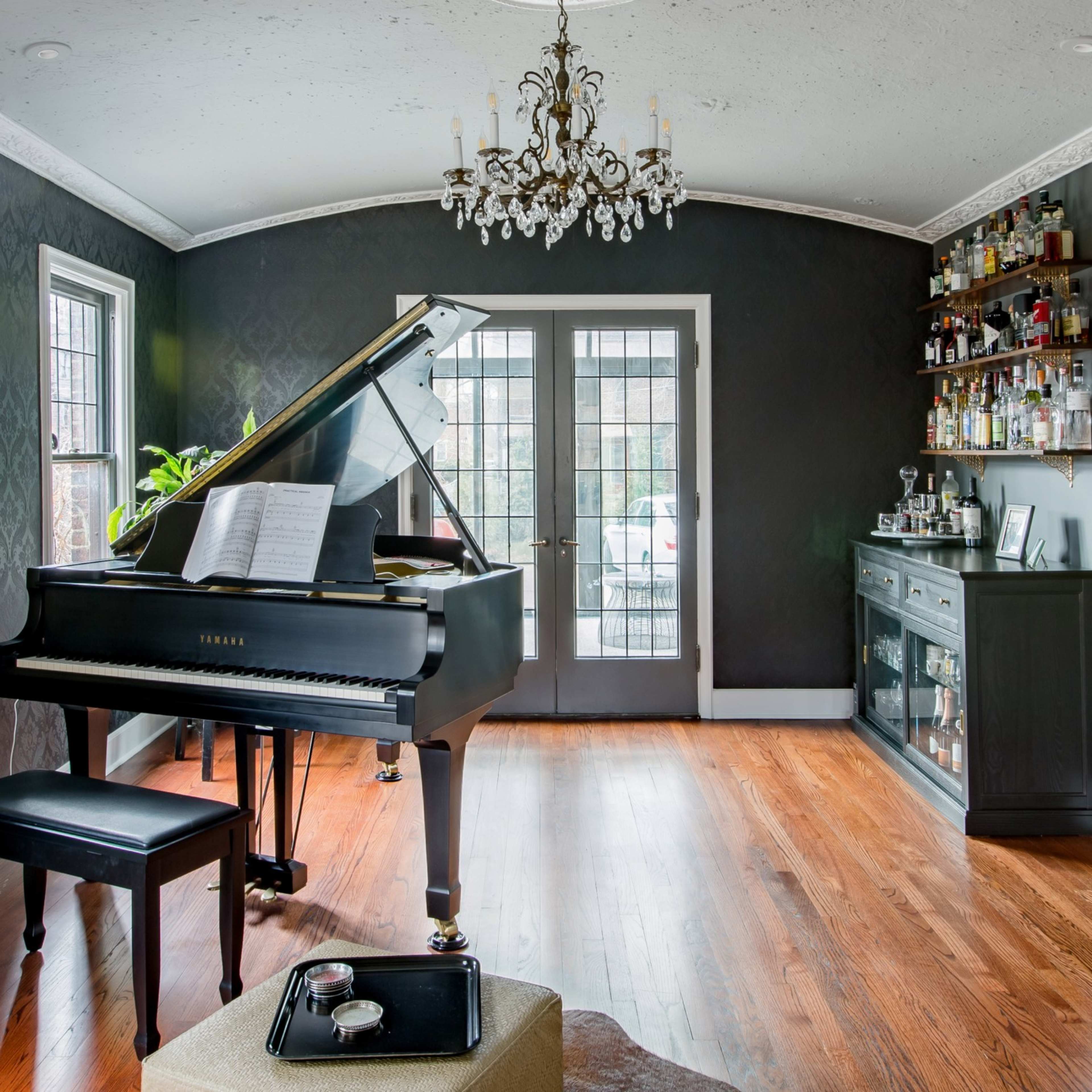A grand piano sits in a stylish room with dark walls, hardwood flooring, and large windows, accompanied by a bar area and dining table.